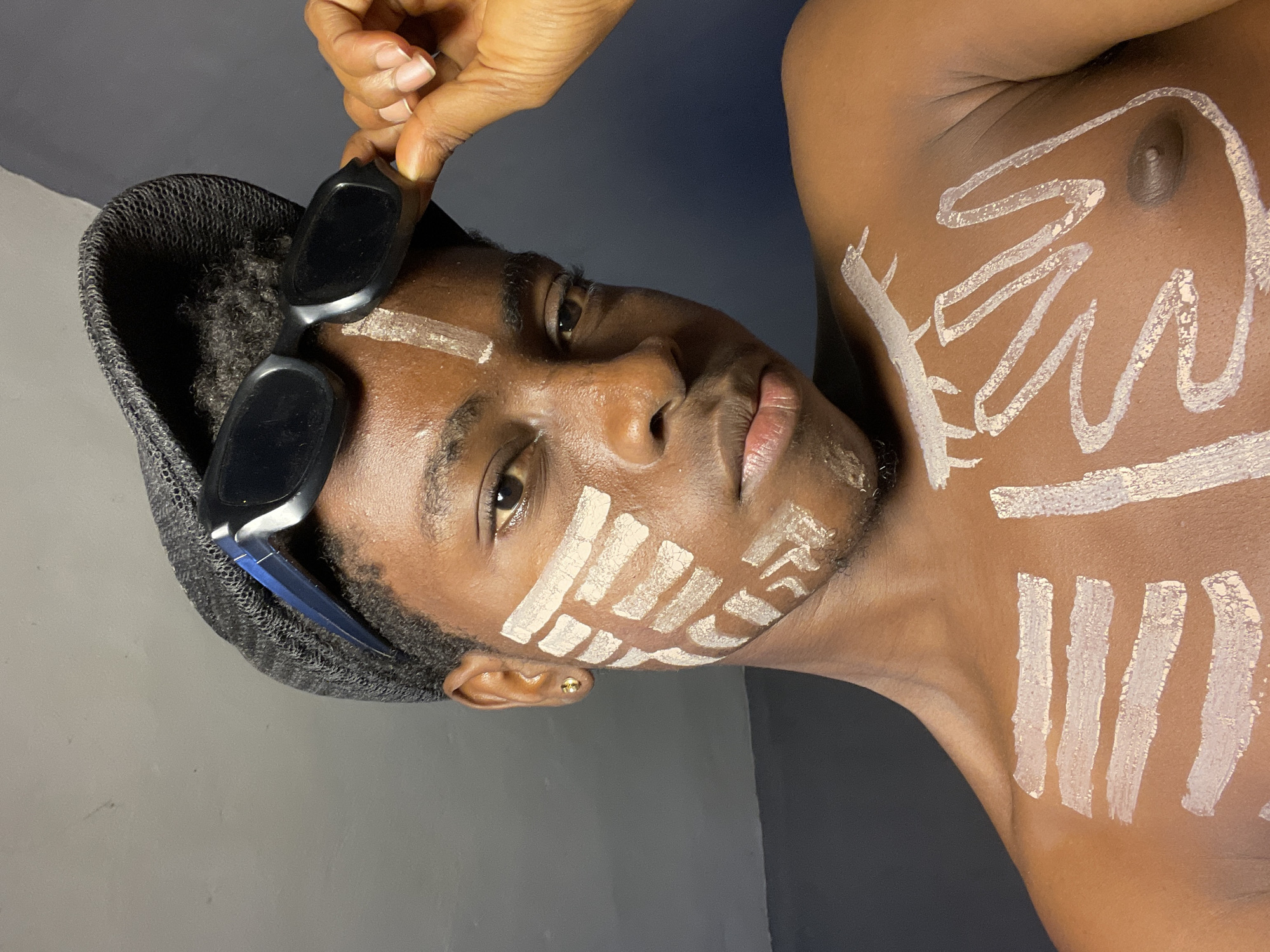 A medium shot portrait shows a dark-skinned person, possibly male, facing slightly left. The person has a hat, sunglasses, and white paint markings on their face and chest. The person is partially nude, with a visible nipple on the right. The hand holding the sunglasses belongs to another person and is positioned in the upper left corner. The background is a plain gray wall. The subject has paint designs on their face and chest, possibly symbolic or decorative. The location, Nkpor, Nigeria, cannot be verified solely from the image.