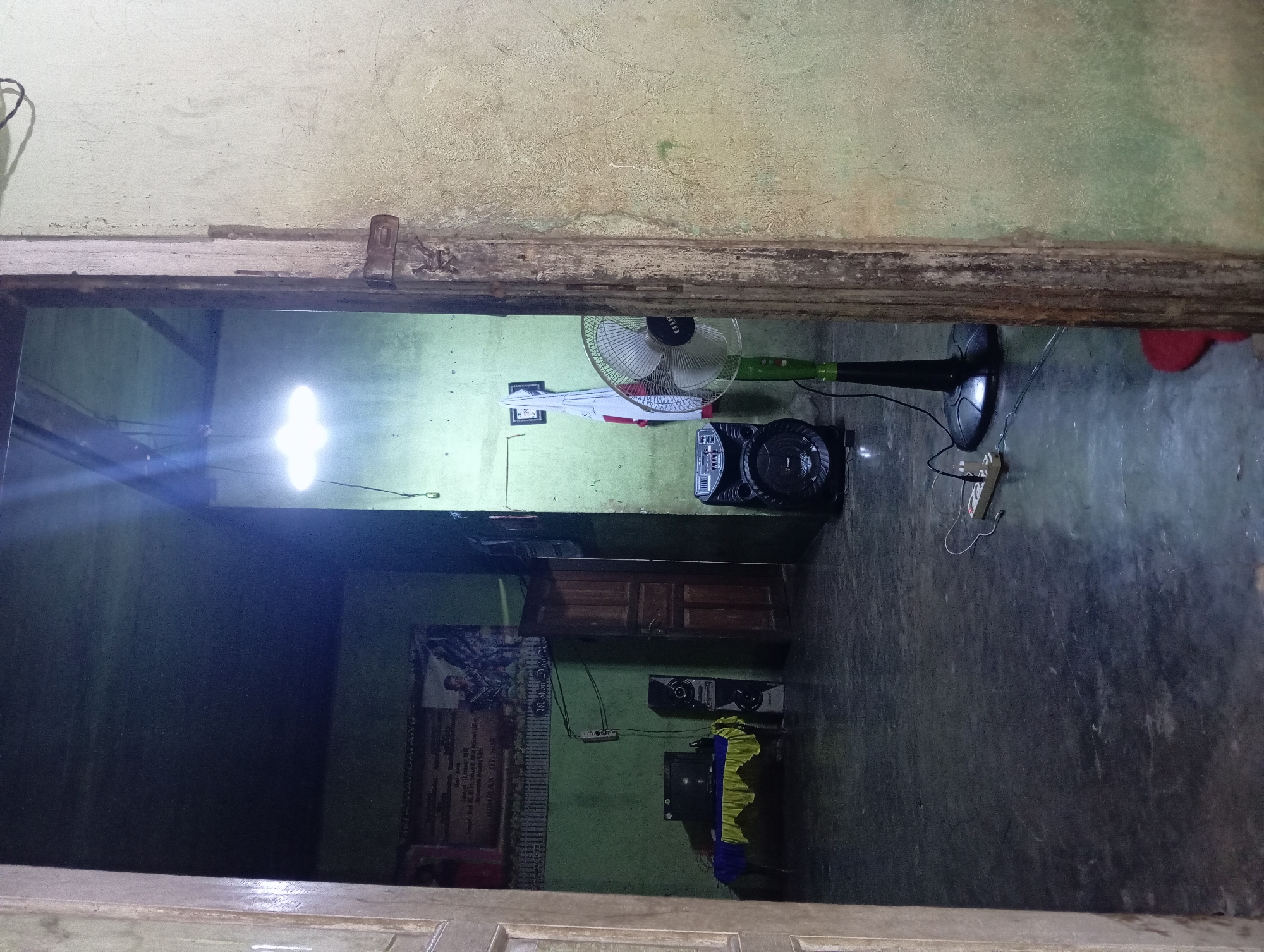 The image shows the interior of a room, possibly in Rejosari, Indonesia. The room appears dimly lit, with a single overhead light source casting a bright glow. The walls are painted a pale green and show signs of wear. A doorway in the foreground frames the view into the room.
Inside, several objects are visible. On the left, a table covered with a yellow and blue cloth holds a television set and what appears to be a bottle. Next to the table are two large speakers. A banner with text and an image is hanging on the wall behind the table.
In the center of the room, a large black speaker is placed on the floor, connected to a power strip with electrical cords. A standing fan with a green pole and black blades is positioned next to the speaker, and a white flag with red accents is draped behind it.
The floor is a dark, smooth surface, possibly concrete or tile. The overall impression is of a lived-in space with basic amenities. The scene suggests a domestic setting, possibly a living area or a common room.