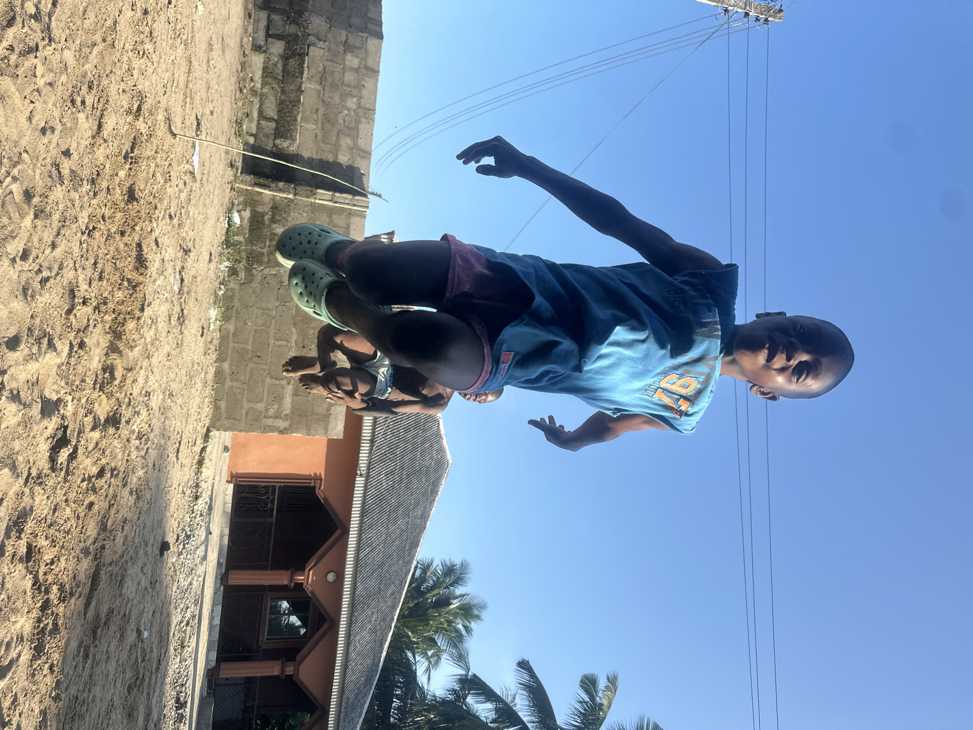 The media file shows two young boys outside in Badagry, Nigeria. The primary subject is a boy in the foreground, wearing a blue t-shirt with orange lettering and pink shorts, and light green Crocs. He is looking directly at the camera with a slight smile. His right arm is outstretched. Behind him, a second boy is visible, wearing a grey t-shirt and black shorts. He is lying down and appears to be holding the first boy's leg.
The scene is a dusty outdoor environment. To the left, there is a sandy ground with some debris. In the background, there is a brick building with a reddish-brown roof, and palm trees are visible. Overhead, a clear blue sky dominates the scene, with power lines stretching across it. The lighting suggests it is daytime, likely late morning or early afternoon, with bright sunlight casting shadows. The overall mood appears relaxed and playful. The number "76" is visible on the t-shirt of the boy in the foreground.