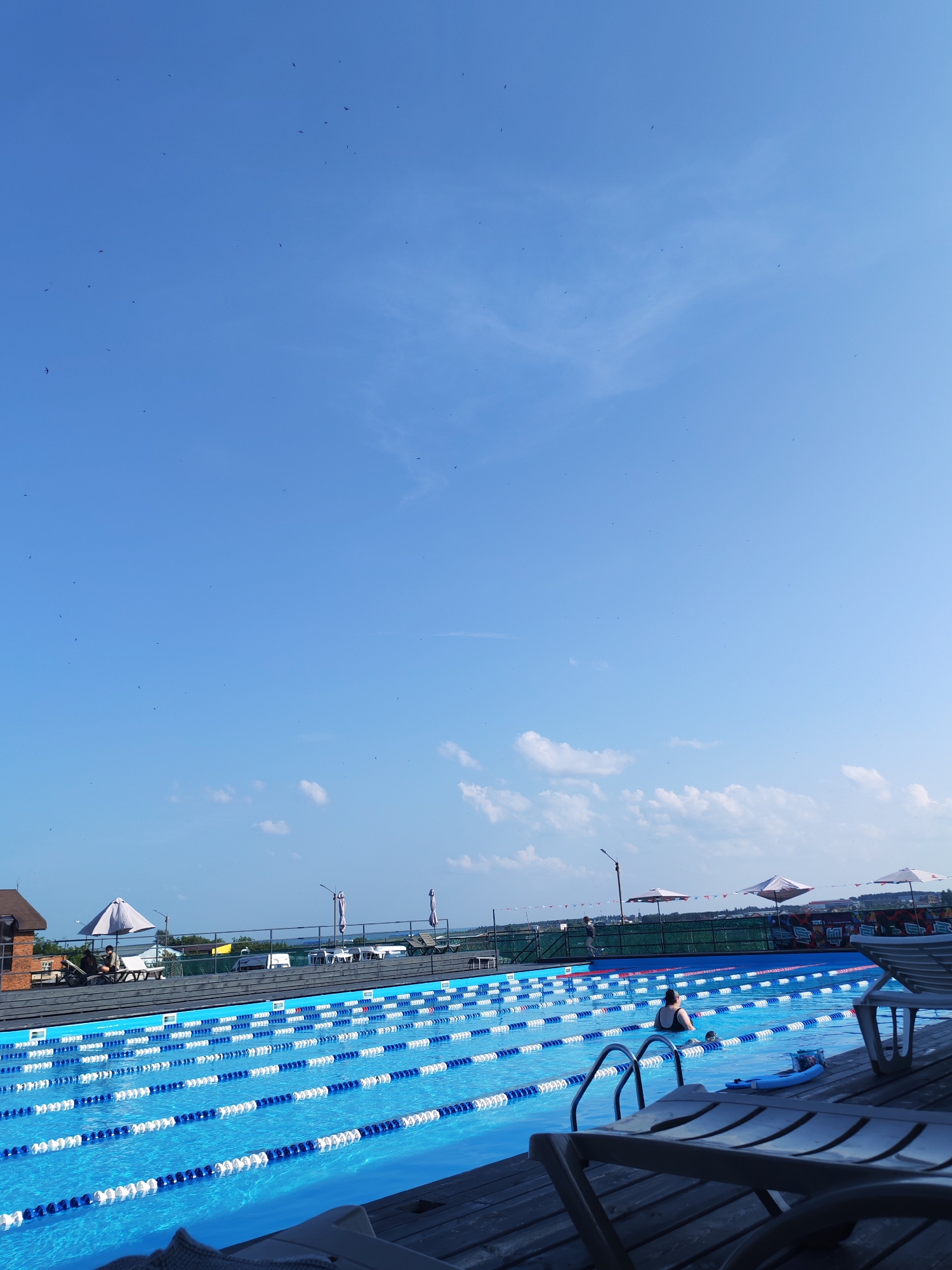 The image captures a bright, sunny day at an outdoor swimming pool facility, possibly located in Pirogovo, Russia, given the architectural style of the building and the overall atmosphere. In the foreground, several lounge chairs are positioned on a wooden deck, some in focus and others blurred. The pool itself is a prominent feature, divided into multiple lanes by blue and white lane ropes. The water is a vibrant blue, indicating it is clean and well-maintained.
In the middle ground, a woman in a dark swimsuit is visible in the pool, appearing to be swimming or resting. Near her, a child is also present in the water. Several people are seated on lounge chairs near the edge of the pool, under large white umbrellas. The background reveals a building with a distinctive brown and red brick facade, reminiscent of Russian architecture. Beyond the pool area, a fence separates it from a grassy area with more umbrellas and some distant structures. The sky is a clear, bright blue with a few wispy clouds scattered across it. Numerous small dark specks are visible in the sky, likely birds in flight. The overall scene suggests a leisurely day at a recreational facility.