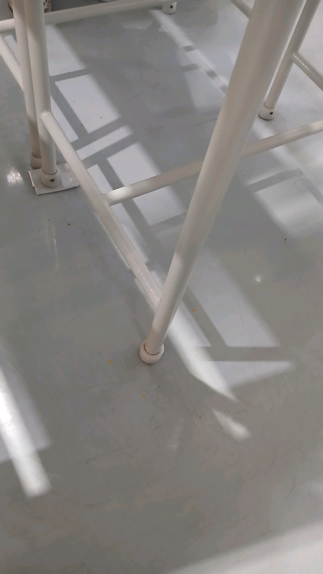 The image depicts a close-up, low-angle view of the white metal frame of a table or chair resting on a light grey, glossy floor. The frame consists of several vertical and horizontal bars. Light casts distinct rectangular and linear shadows across the floor, originating from the overhead structure of the furniture. There are a few small dark specks and some minor scuff marks visible on the floor's surface. One of the legs of the furniture has a white circular cap at its base. The scene is indoors, likely in an industrial or institutional setting, given the utilitarian nature of the furniture and the seamless flooring. There is no discernible indication of the location being in Gadobangkong, Indonesia, nor are there any people or specific actions depicted.