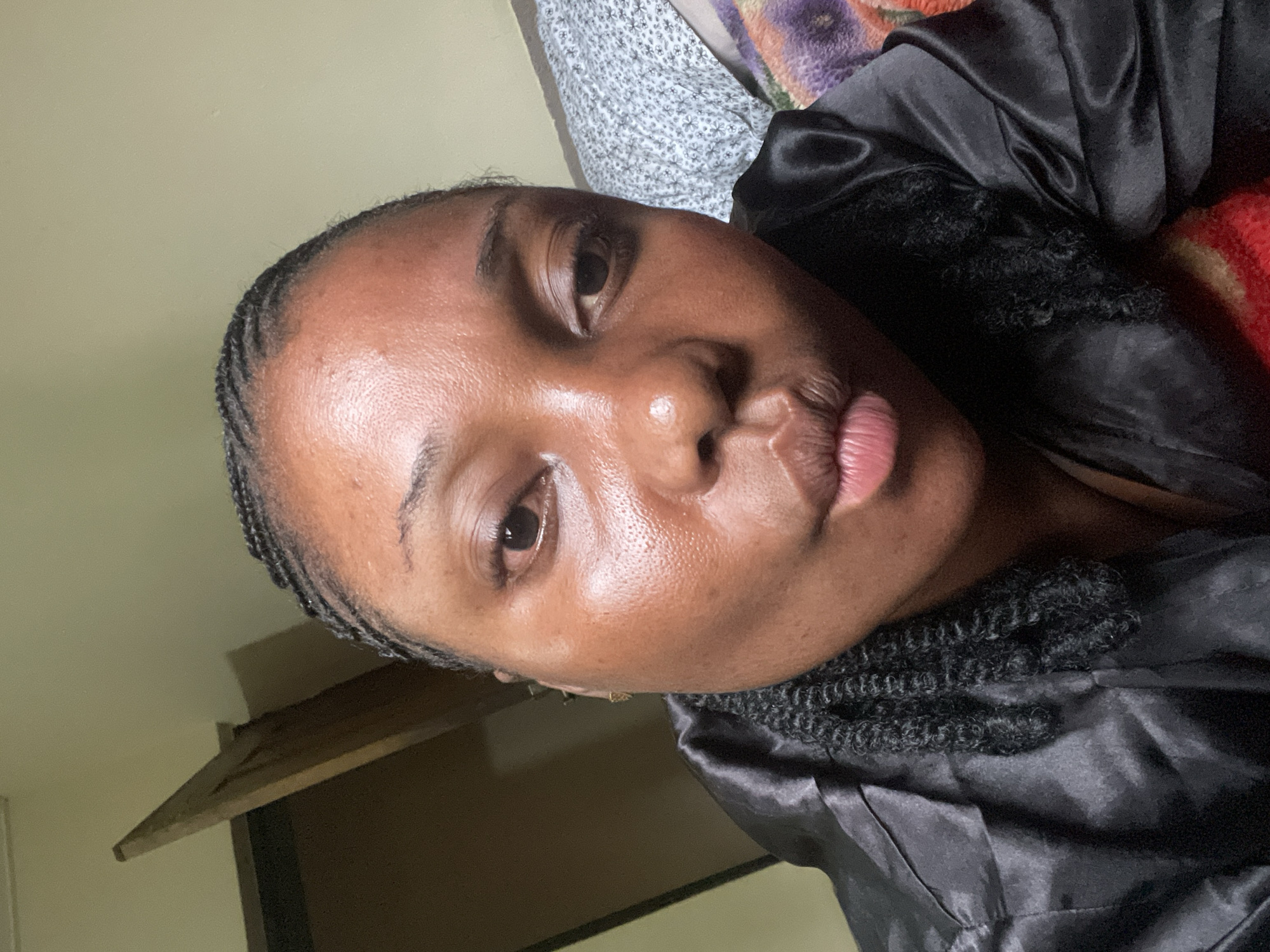 The image is a close-up, rotated selfie of a young adult individual, likely female, from Jos, Nigeria. The person has dark skin and is looking directly at the camera with a playful "kiss face" expression, featuring pursed lips. Their dark hair is styled in braids, with cornrows visible along the hairline and looser, curly braids around the neck and shoulder area.
They are wearing a black satin or silky garment, possibly a robe or top, which drapes softly and reflects light. The setting is indoors, appearing to be a bedroom or personal space. Behind the person's head, to the left, there's a light grey or white patterned fabric, possibly a blanket or sheet. To the right, a colorful knitted or woven blanket with shades of red, orange, and purple is visible. The background also shows a plain, light-colored wall (beige/greenish-yellow) and a section of what looks like wooden furniture on the lower left. The lighting is soft and ambient, suggesting an indoor environment during evening or night. No text is visible in the image. The overall mood is casual and expressive.