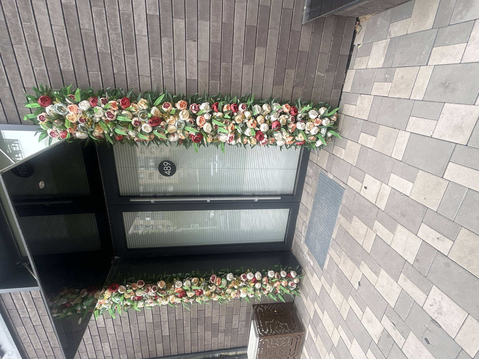 This is a full-shot, eye-level, exterior photograph of a building entrance in Almaty, Kazakhstan. The entrance features double glass doors framed by tall, vertical arrangements of artificial flowers in shades of red, pink, cream, and green. A black awning extends over the doors. The building's facade is clad in dark gray brick. To the left of the entrance, a metallic square planter with decorative cutouts sits on the patterned gray and beige tiled sidewalk. A circular metal grate is embedded in the sidewalk in front of the doors. The number "489" is displayed on a circular black plaque on the left door. The scene is well-lit, suggesting daytime.