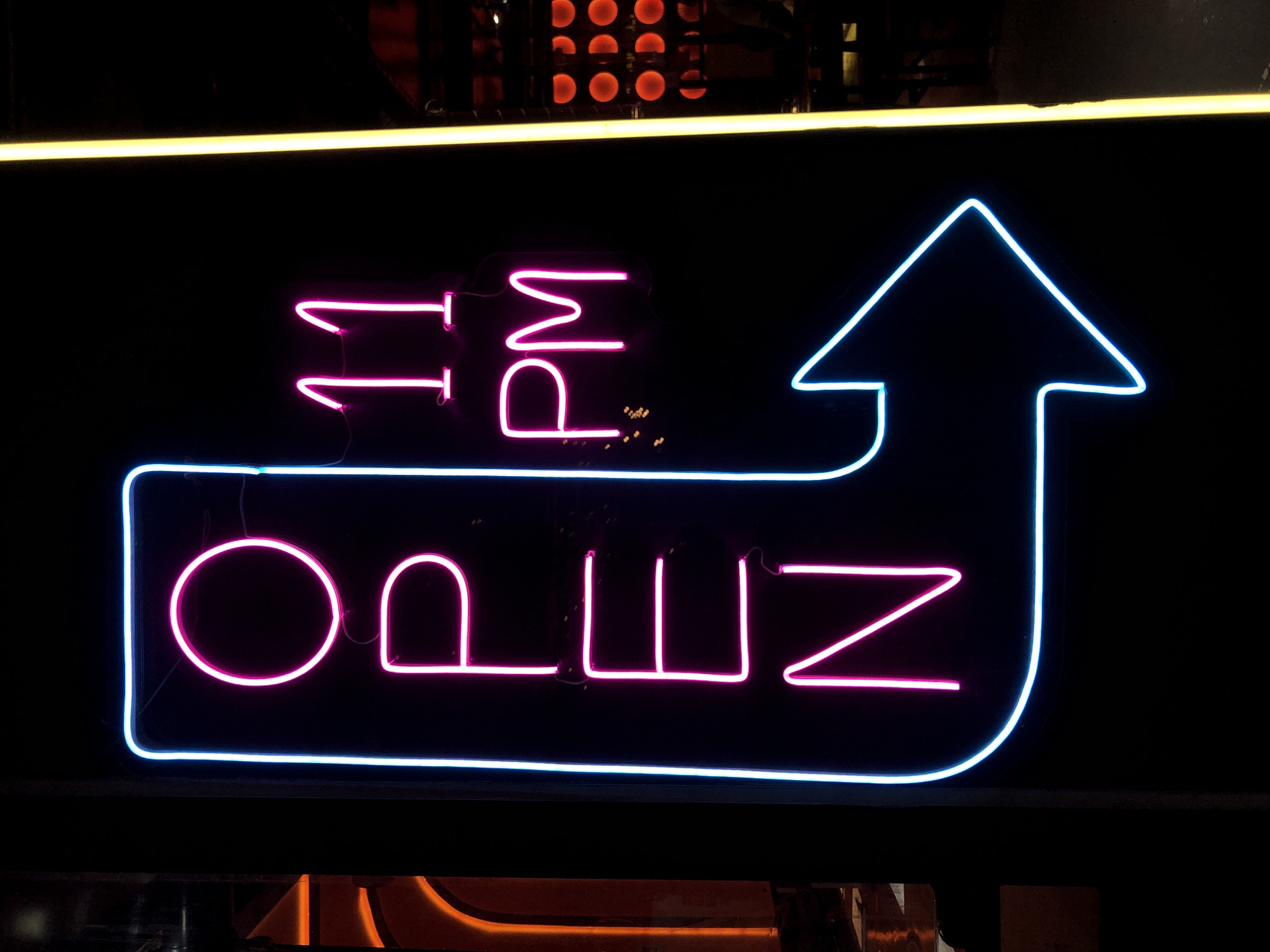 This is a close-up, eye-level shot of a neon sign that reads "OPEN 11 PM" with an upward-pointing arrow. The sign is predominantly pink and blue, with the word "OPEN" in pink and the "11 PM" and arrow in pink and blue neon respectively. The sign is set against a dark background, possibly a storefront or building facade. A yellow neon strip runs horizontally above the "11 PM" portion of the sign. The overall impression is one of nighttime ambiance, likely in a commercial or entertainment setting. The scene appears to be outdoors, given the reflection of what looks like streetlights or building lights at the bottom of the image. There are no people visible in the frame. The neon lights are vibrant and clearly illuminate the text and the arrow, indicating that the establishment is open for business. The location context is Đà Nẵng, Vietnam, which suggests a potentially bustling urban environment. The specific time is indicated by the "11 PM" text.