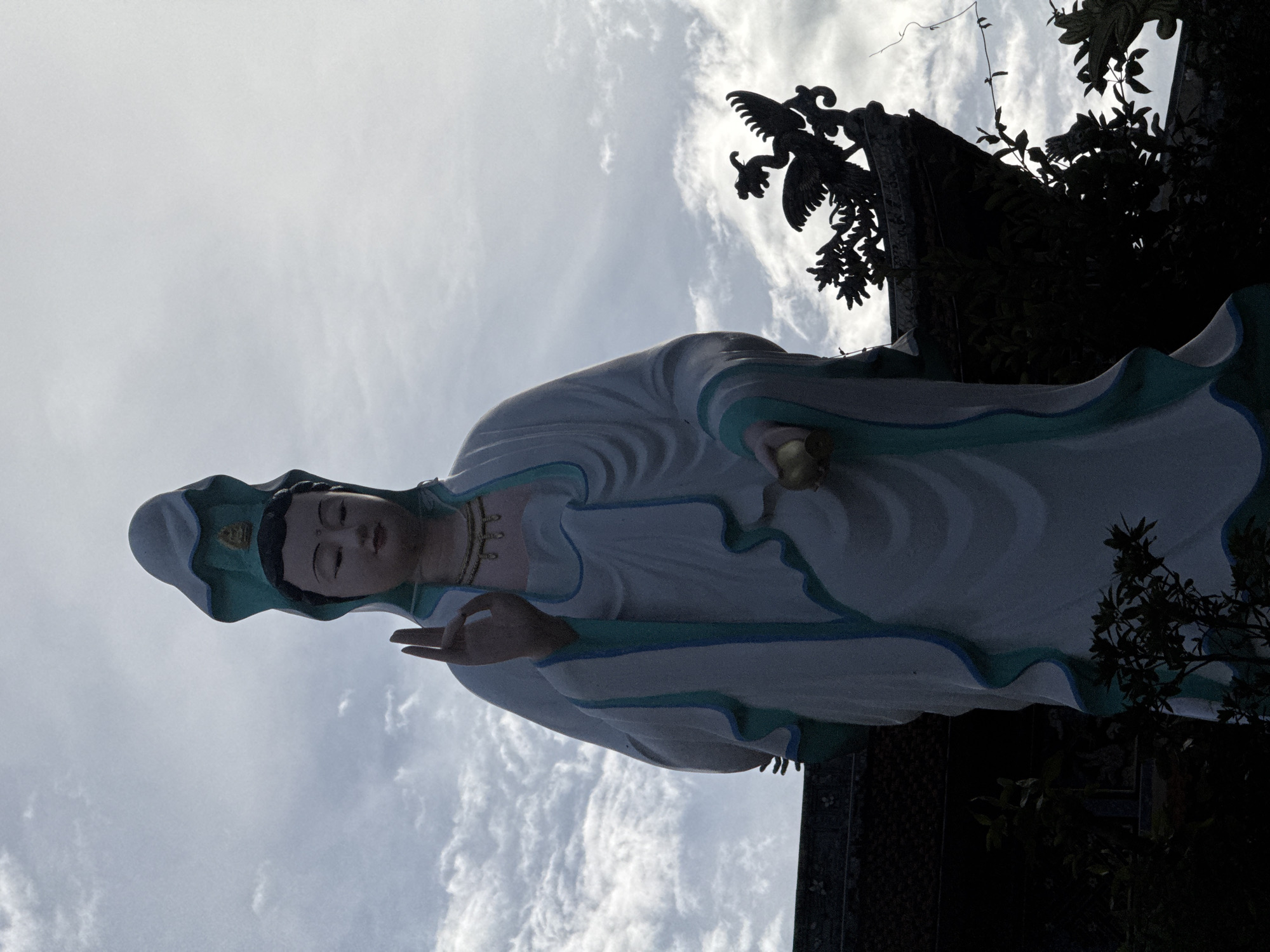 A full-body, low-angle shot captures a large, white statue of a female deity in traditional robes, possibly Guanyin, against a cloudy sky. The statue has a serene expression, wears a green and white headpiece, and holds a small, golden orb in its left hand. Its right hand is raised with fingers pointing upwards in a gesture. The statue is adorned with a gold necklace. In the foreground and lower right of the image, dark silhouettes of foliage and intricate architectural elements, including a decorative roofline topped with a phoenix figurine, are visible. These architectural details suggest the statue is located in a temple or similar religious site, possibly in Da Lat, Vietnam, known for its many pagodas and spiritual landmarks. The overall mood is solemn and awe-inspiring, with the natural light illuminating the statue and the sky.