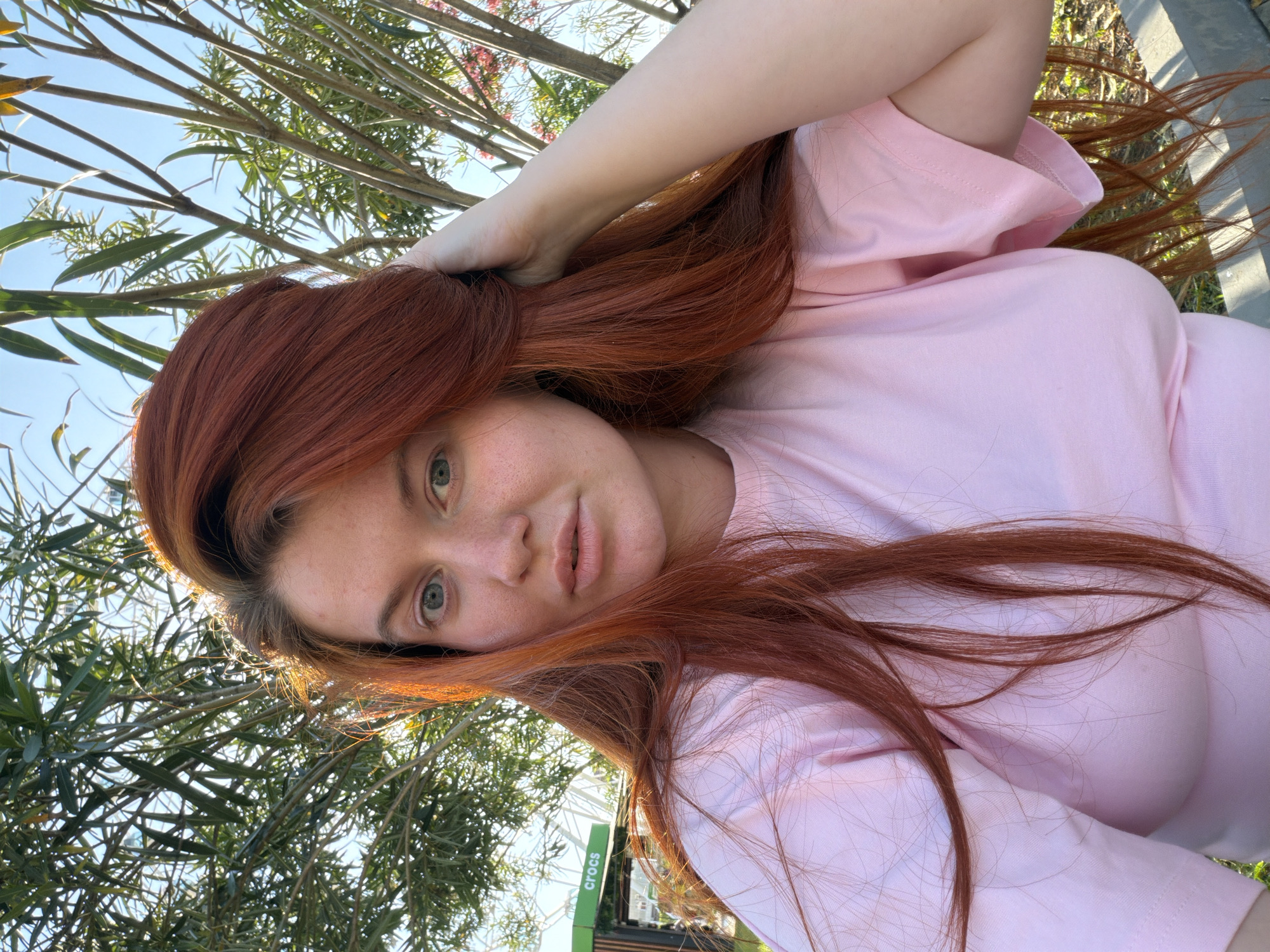 A young woman with long, reddish-brown hair and light green eyes is captured in a close-up, sideways selfie. She is wearing a pink t-shirt and is outdoors, with lush green foliage and a bright blue sky visible in the background. Her hand is resting on her head, pushing her hair back. The lighting suggests it is daytime, likely sunny, with some shadows indicating the sun's position. A partially visible sign with the word "Crocs" is present in the lower right portion of the image. The overall impression is casual and relaxed, possibly taken on a warm, bright day.