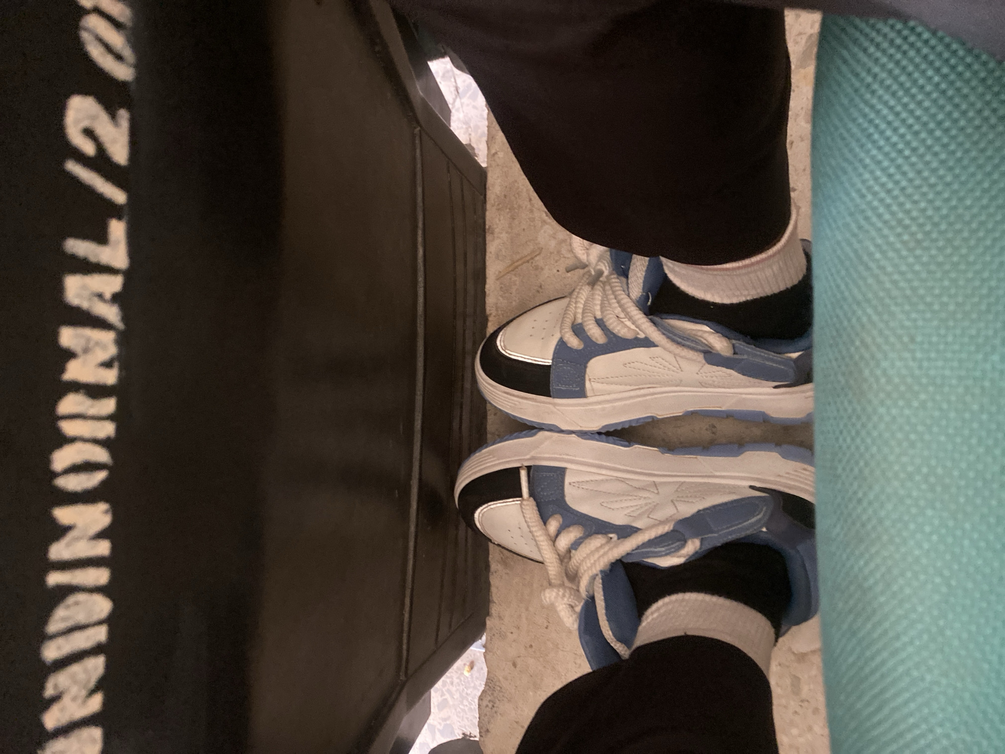 The image shows a pair of blue and white sneakers with white laces, worn by a person sitting on a turquoise surface. The person is wearing black pants and white socks. Above the person's feet, there is a dark-colored object with white lettering that appears to read "NIDINORMAL/2 0". The background is a concrete surface. The scene appears to be indoors or in a sheltered outdoor area. There are no specific visual cues to pinpoint the location to Jalingo, Nigeria.
