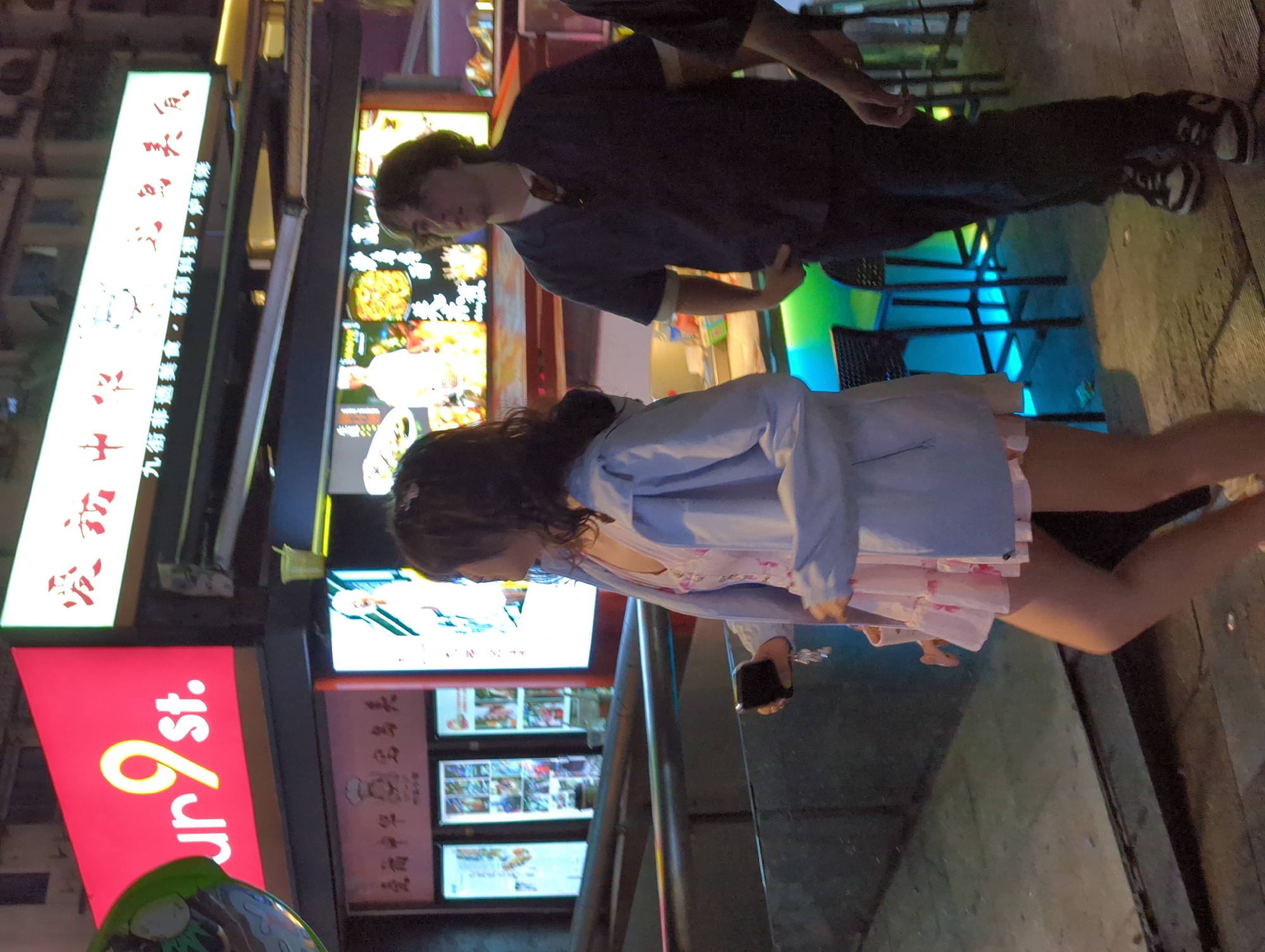 The photograph captures an outdoor scene featuring two individuals, along with the signage of a business named "9st." and Chinese writing. A person with dark hair, dressed in a light blue jacket and a floral patterned dress, appears to be using a mobile phone. Next to them stands another individual with light hair, donning a dark blue shirt and dark pants, completing their look with a pair of sneakers. The setting seems to be a bustling street, with a storefront display behind them, exhibiting several menu options and other promotional content. To the left, a red signboard bearing the name "9st." is visible. Additional signage in Chinese script suggests a dining or commercial establishment nearby. The photograph presents a dynamic interplay of individuals within an urban landscape marked by commerce and signage. It is impossible to determine if this photograph was taken in the Liangjiang area of China without additional information.