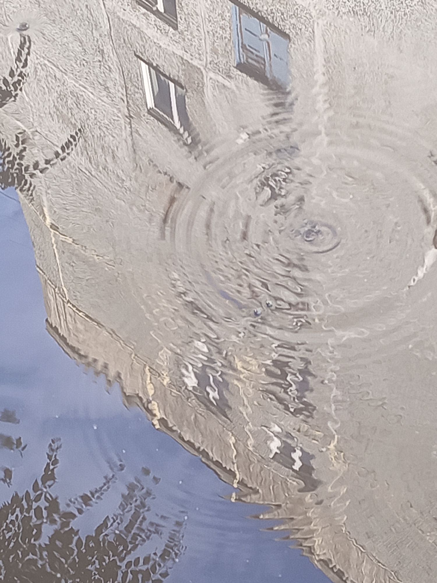 The image depicts a reflection in a body of water, likely a puddle, showing a distorted view of a building and tree branches. The building appears to be made of concrete or a similar material and has several windows. The water's surface is disturbed by ripples, suggesting recent rainfall or a disturbance. The scene is rendered in a soft, painterly style, with muted colors. The location is identified as Novokuznetsk, Russia. There are no people visible in the image. The context implies an urban environment experiencing wet weather.
