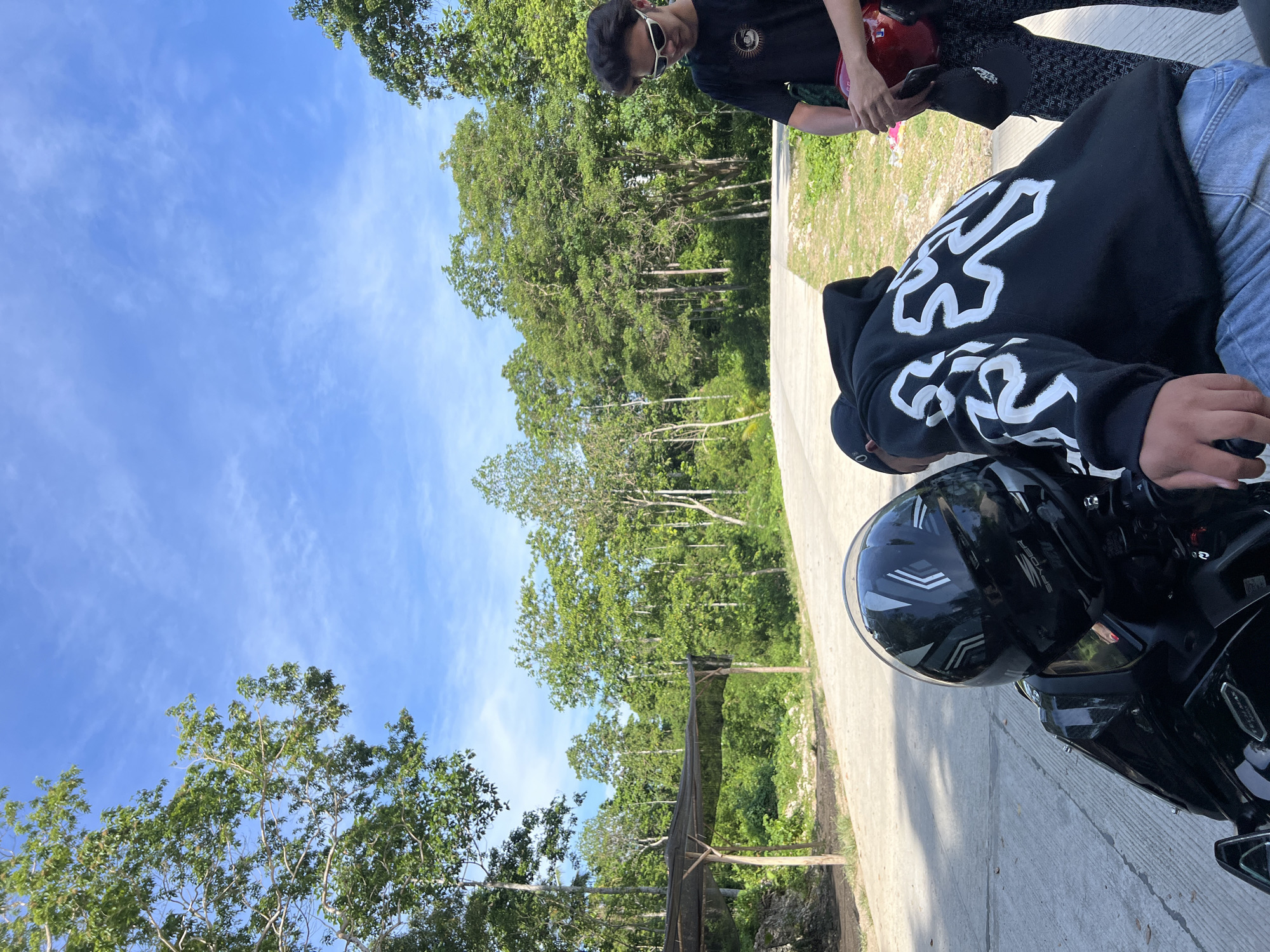 In Samal, Philippines, two individuals are captured in an outdoor scene with a focus on a motorcycle and its rider. The rider, wearing a black hoodie with a large white cross design and blue jeans, is leaning forward on the motorcycle, his face obscured. A black motorcycle helmet with silver accents sits on the motorcycle's seat. To the right, another person stands, wearing a black t-shirt, sunglasses, and patterned pants, holding a red helmet. The background features a concrete road, lush green trees, and a clear blue sky with wispy clouds, suggesting a bright, sunny day. A simple open shelter with a black mesh roof is also visible in the mid-ground.