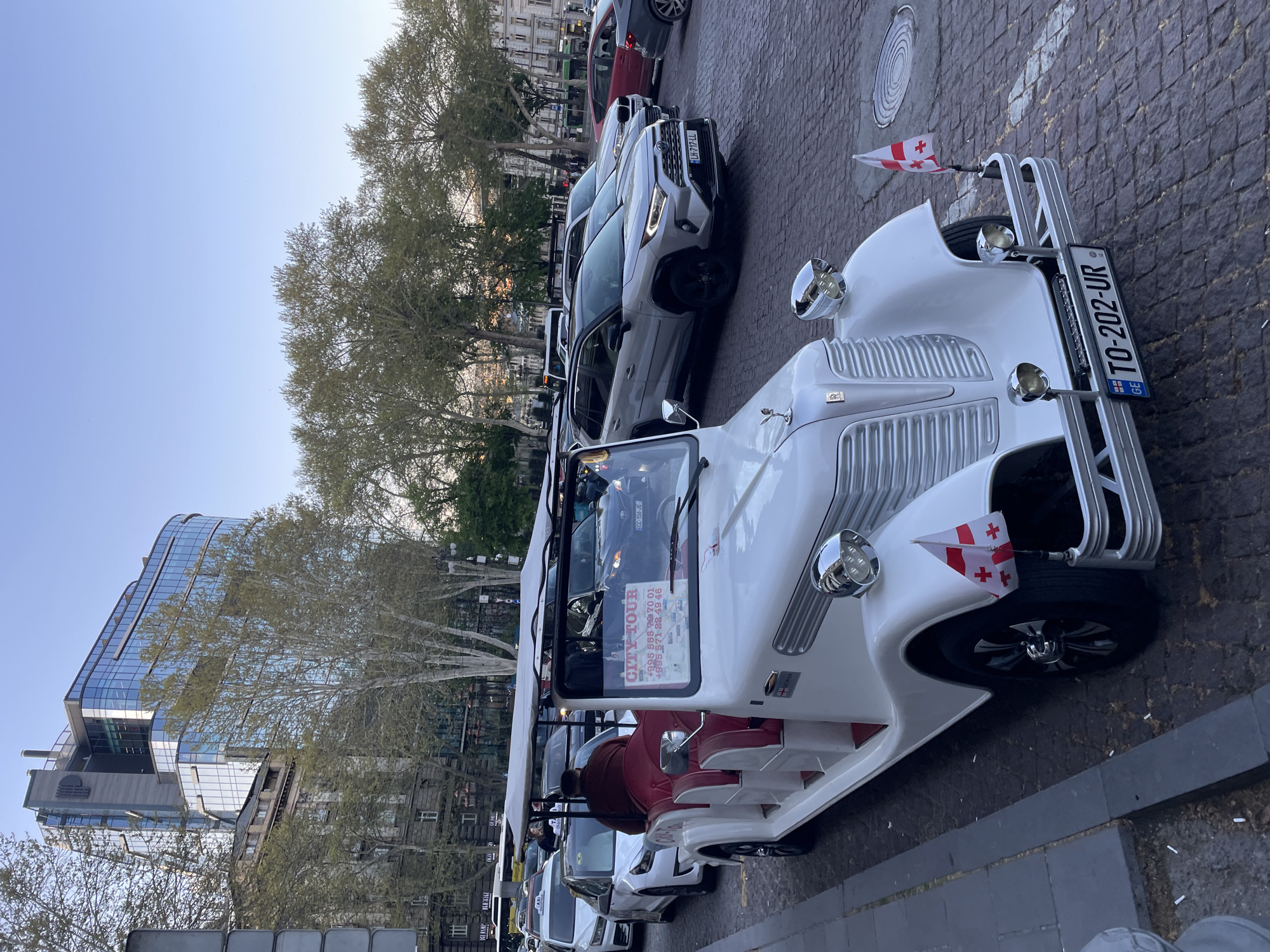 An eye-level outdoor shot captures a white, vintage-style city tour vehicle parked on a cobblestone street in Tbilisi, Georgia. The vehicle, likely an electric or modified car, features a distinctive front grille, shiny chrome headlights, and a red interior visible through the open top. Small Georgian flags are mounted on the front fenders. A person wearing a red top is visible in the driver's seat.
Behind the tour car, several modern cars, including SUVs and sedans, are parked in a line. Trees with sparse, light green foliage line the street, suggesting either early spring or late autumn. In the background, a contemporary glass-fronted building stands tall next to older, more traditional urban architecture, reflecting a mix of old and new common in the city. The sky above is clear and light blue, indicating daytime with good weather.
Visible text on a sign in the tour car's window reads "CITY TOUR" along with two phone numbers: "+995 555 79 70 01" and "+995 571 22 49 46". The Georgian license plate on the front of the tour car displays "TO-202-UR" and the country code "GE". Another license plate on a car behind it shows "LH-717-HL". Partially visible text on a distant building spells "BLANC".