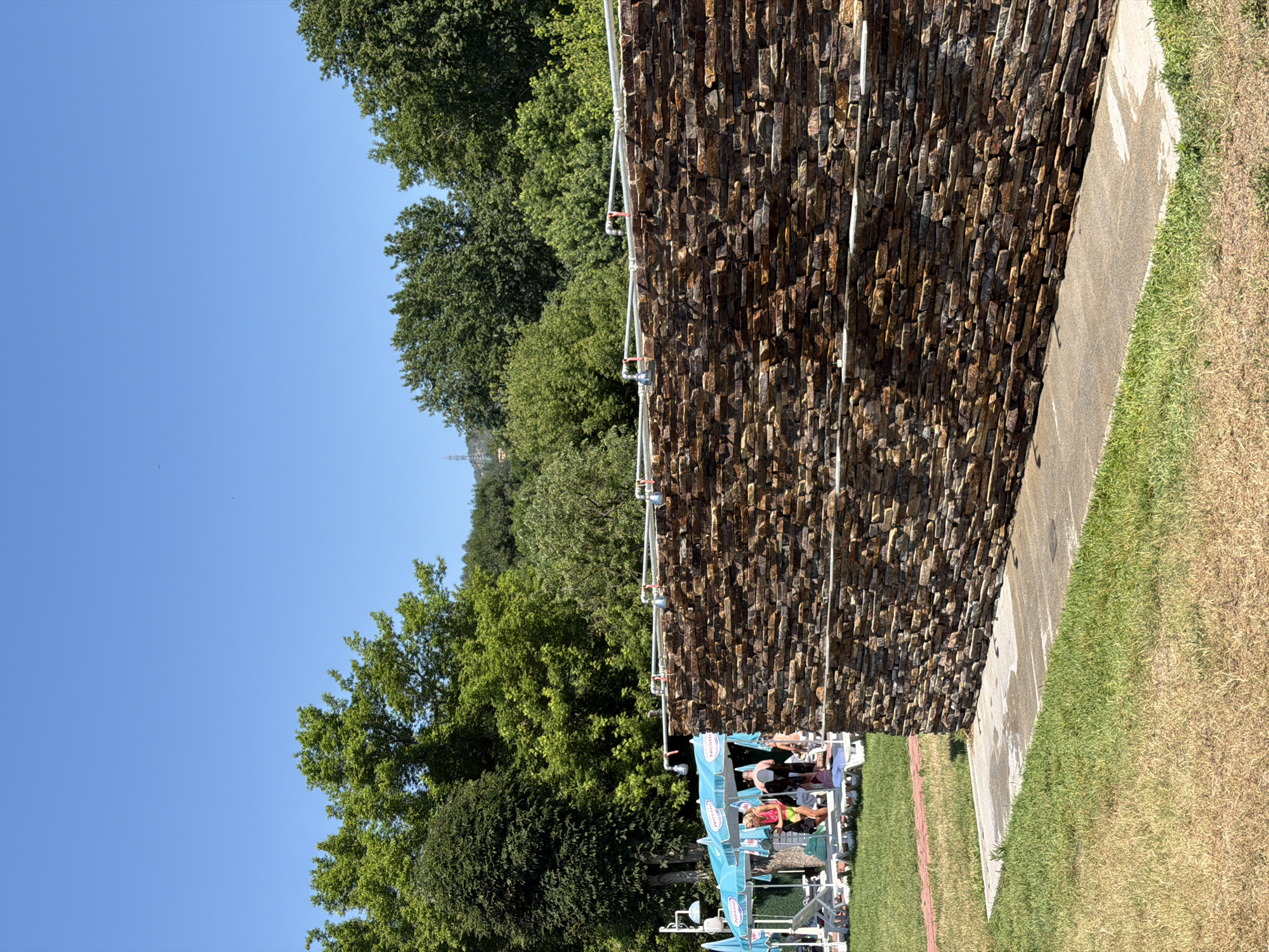 The image depicts an outdoor scene on a sunny day, likely at a recreational area in Simitli, Bulgaria, given the context of the provided crops. In the foreground, a textured stone wall, constructed from horizontally stacked rectangular stones in varying shades of brown and orange, dominates the right side of the frame. Water pipes with visible faucets run along the top of this wall, suggesting it may be part of an outdoor shower or washing facility.
To the left of the stone wall, a patch of concrete is visible, wet in places, indicating recent water usage. Beyond the concrete, green grass covers the ground, transitioning to drier, yellowed grass further into the foreground. A red brick path is partially visible in the middle ground, leading towards a shaded area.
Underneath several blue umbrellas with "Nestle" branding, lounge chairs are set up on the grass. Several people are present in this area. One person, wearing a pink top and yellow shorts, stands near the chairs. Another person in a black swimsuit is also visible. Other individuals are present but less clearly defined. In the background, a dense line of lush green trees forms a natural backdrop, with a hazy outline of hills or mountains visible in the far distance under a clear blue sky. The overall impression is of a summer day at an outdoor facility, possibly a park or a resort, with amenities for cooling off.