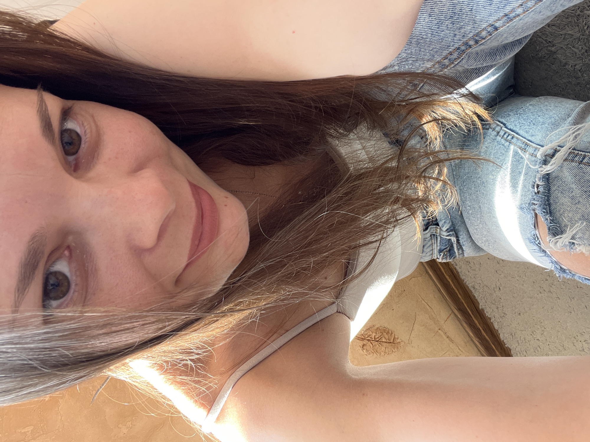 A young woman is captured in a selfie, the image oriented vertically with the camera looking down. She has long, dark brown hair that cascades around her face and shoulders. Her eyes are a warm brown, and she has a slight smile on her lips, conveying a relaxed and possibly content emotion. She is wearing a light-colored tank top and distressed denim shorts. The background is a mix of textures and colors, with what appears to be a wooden floor or surface in the foreground and a carpeted or textured area in the background, along with what looks like a wall. Sunlight is streaming in, creating bright highlights on her skin and hair, suggesting it is daytime and likely sunny. The overall setting appears to be indoors, perhaps a bedroom or living space, given the indoor textures and natural light. There are no visible text elements in the image. The context provided indicates the location is Tolyatti, Russia.