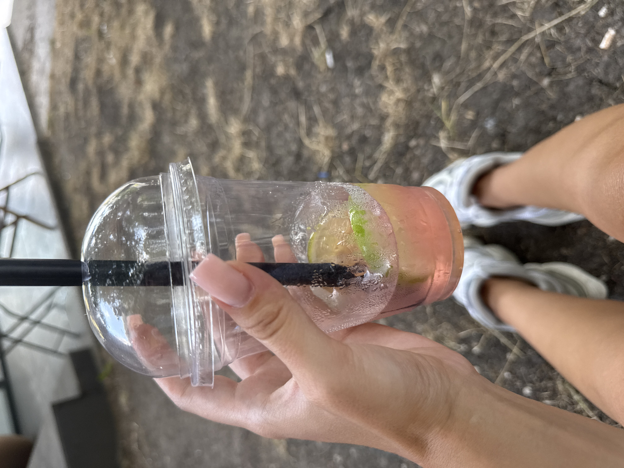 In Chișinău, Moldova, a person's hand, with neatly manicured pink nails, holds a clear plastic cup containing a pink beverage with slices of citrus fruit and possibly mint. A black straw extends from the cup. The cup is sweating, indicating it contains a cold drink. The background suggests an outdoor setting with dry, scattered vegetation and what appears to be the legs and feet of the person, wearing white sneakers. The overall scene implies a casual, relaxed moment, possibly at an outdoor cafe or park.