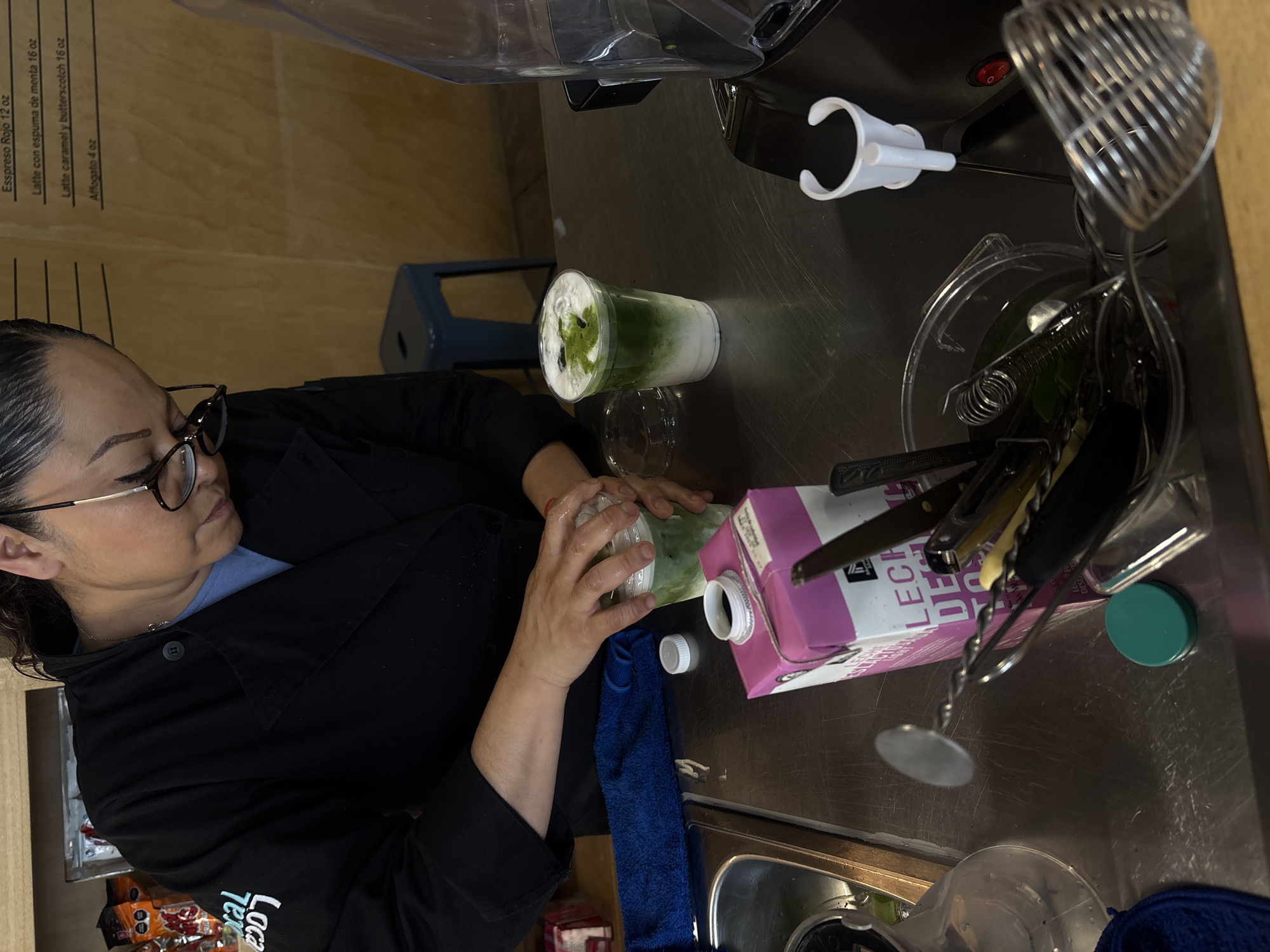 The image shows a woman wearing glasses and a black jacket with a logo, standing behind a metal countertop. She is in the process of preparing a drink, possibly matcha, with ingredients and equipment nearby including a carton of "Leche Desl" (skim milk), a blender, and various utensils. The background features a wooden wall with a menu visible in the top left corner, listing espresso, latte, and affogato options. Based on the menu in the background, the location is likely in Mexico City, Mexico.