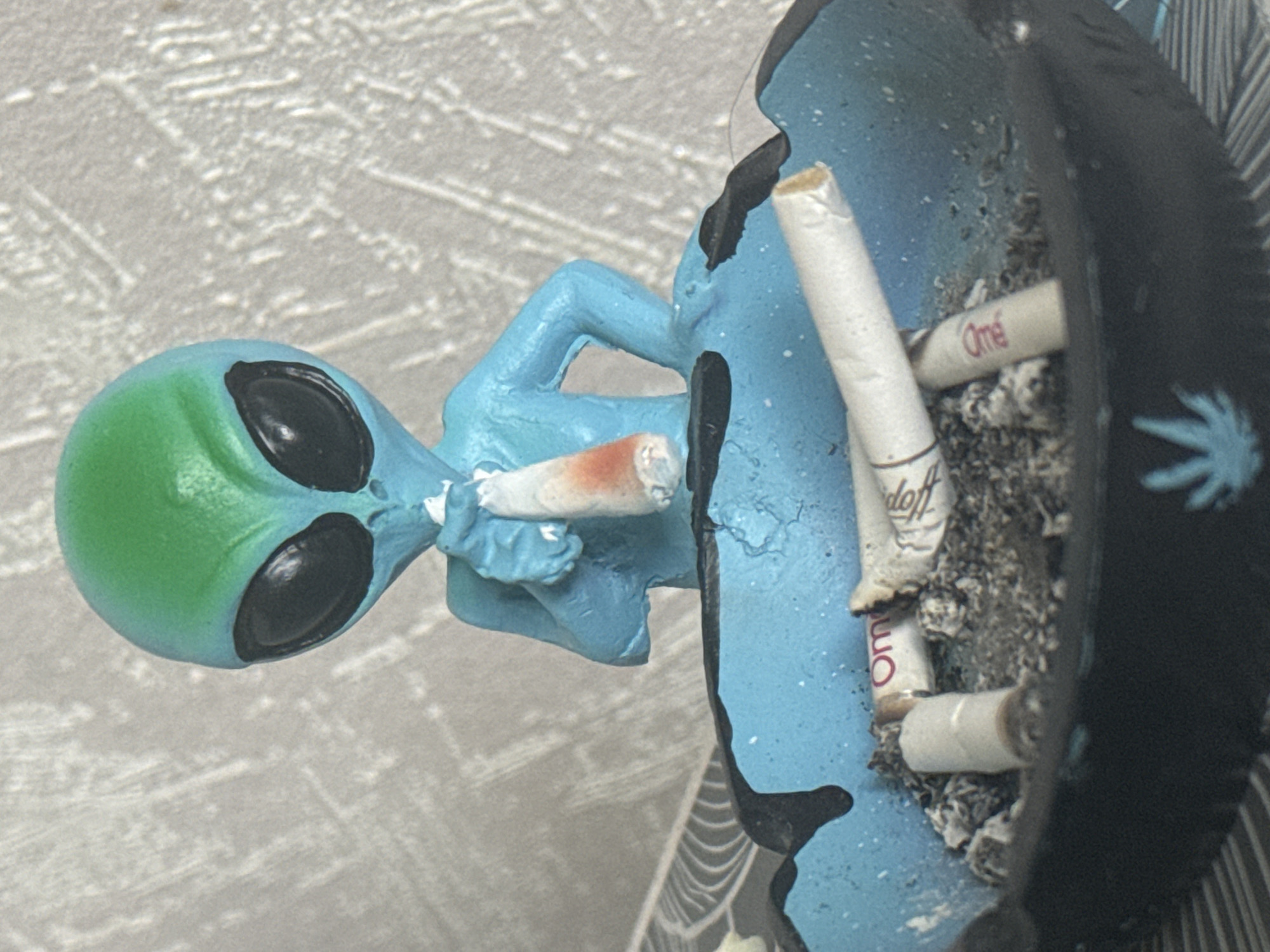 The image depicts a close-up, slightly angled shot of a novelty ashtray shaped like a blue and green alien figure. The alien is holding a lit cigarette to its mouth. The ashtray itself is shallow, blue, and appears to be made of resin or a similar material. It contains several cigarette butts and ash. The background is a textured, light-colored surface. The image's lighting is somewhat harsh, creating noticeable shadows. There is no discernible location in Sofia, Bulgaria, or any other specific geographical context provided by the visual elements.