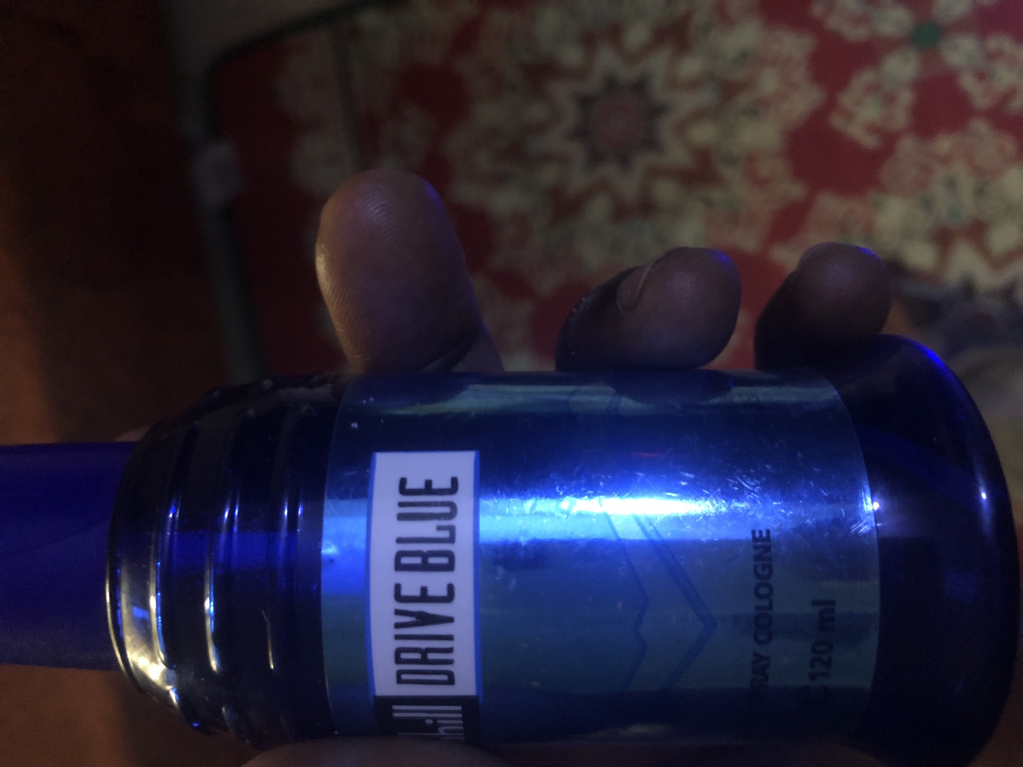 A close-up, overhead shot shows a blue bottle of "DRIVE BLUE" spray cologne held by a person's hand. The bottle is cylindrical with a ribbed cap. The label on the bottle is white with black text. Below the product name, the text "SPRAY COLOGNE" is visible, followed by "120 ml". The background is out of focus, showing a patterned surface in red and yellow hues. The overall lighting is dim, with a strong blue reflection from the bottle. The image was taken in Kabong, Nigeria.