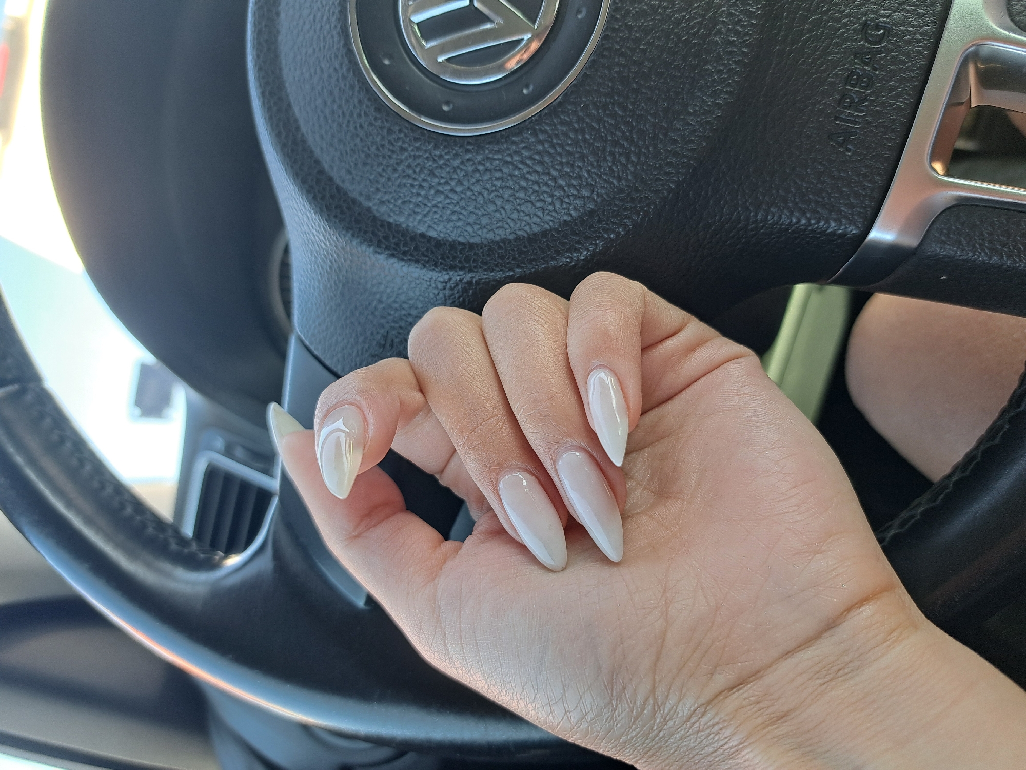 A hand with light-colored, pointed fingernails is resting on a black leather steering wheel. The steering wheel has a silver trim and a Volkswagen logo in the center. The background is blurred but appears to be inside a car, possibly with a view of the outside through a windshield. The image is taken from a close-up perspective, focusing on the hand and the steering wheel. The lighting suggests it is daytime.