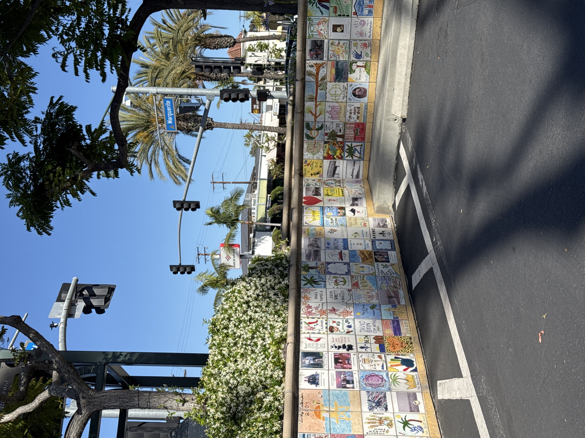 The image shows a section of the tile wall located along Marguerite Avenue in Newport Beach, California. The tiles appear to be individually decorated, possibly by community members or children, with various drawings and names. The wall runs adjacent to a parking area marked with white lines. Above the wall, there is foliage, including white flowering bushes and palm trees. A street sign for Marguerite Avenue is visible, along with traffic lights. The sky is clear and blue, suggesting a sunny day. The image is oriented so that the street and sky are above the tile wall. A Bank of America sign is visible in the background.