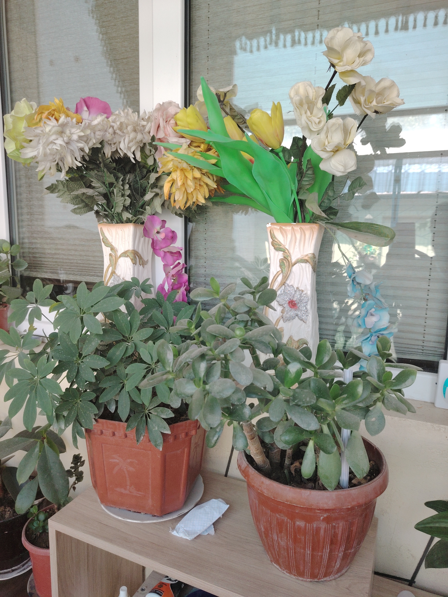 In the city of Unknown, Unknown, a collection of artificial flowers and houseplants is displayed on a shelf in front of a window with blinds. The artificial flowers are arranged in two decorative vases. The vase on the left contains a bouquet of white, pink, and yellow flowers, including what appear to be chrysanthemums and roses. The vase on the right features white roses and yellow tulips, with some green artificial foliage. In the foreground, two potted houseplants are visible. The plant on the left, in a brown square pot with a palm tree relief, has large, compound green leaves. The plant on the right, in a brown oval pot, appears to be a jade plant with thick, succulent leaves. A small, crumpled white paper item rests on the shelf between the two pots. The overall scene suggests an indoor setting, possibly a sunroom or patio, with natural light filtering through the blinds.