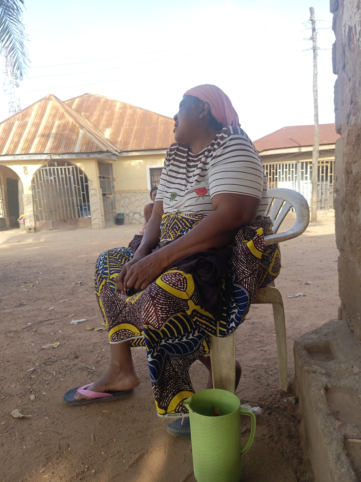 Akwanga, Nigeria. An elderly woman wearing a striped shirt, colorful wrap, and pink headscarf is seated on a white plastic chair outdoors. She looks off to the left of the frame. Her hands rest in her lap. She wears a pair of flip-flops, one black and one black with a pink strap. Next to the chair is a green plastic pitcher on the dirt ground. Behind her, a yellow building with a corrugated metal roof is visible, as well as parts of another building. A portion of a dilapidated wall is on the right side of the frame. The ground is dirt with scattered debris.