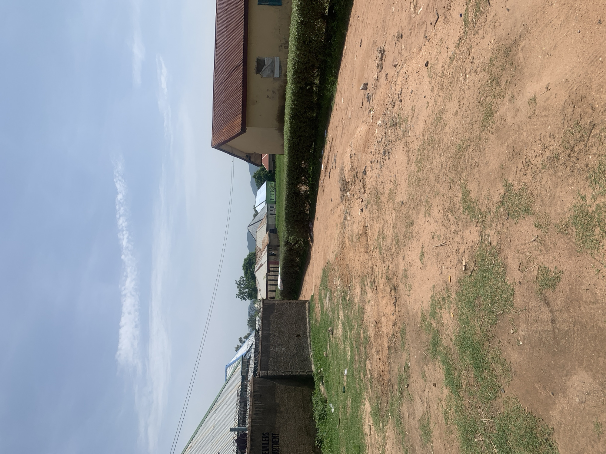 A low-angle, full outdoor shot shows a dirt road leading through a rural area. On the left, a concrete wall with barbed wire on top runs along the edge of the road. The wall has text that appears to read "EVAILERS APARTMENT." Beyond the wall, a corrugated metal roof of a building is visible. On the right side of the road, a neatly trimmed green hedge runs parallel to the path. Behind the hedge, a yellow building with a rusty corrugated metal roof stands. Several other low-rise buildings with similar roofs are visible in the distance, some painted in pastel colors. The sky is clear blue with a few wispy white clouds. The ground is covered in reddish-brown dirt with patches of sparse green grass. The overall impression is of a simple, somewhat weathered community in a dry climate. The location is identified as Jauro Yinu, Nigeria.