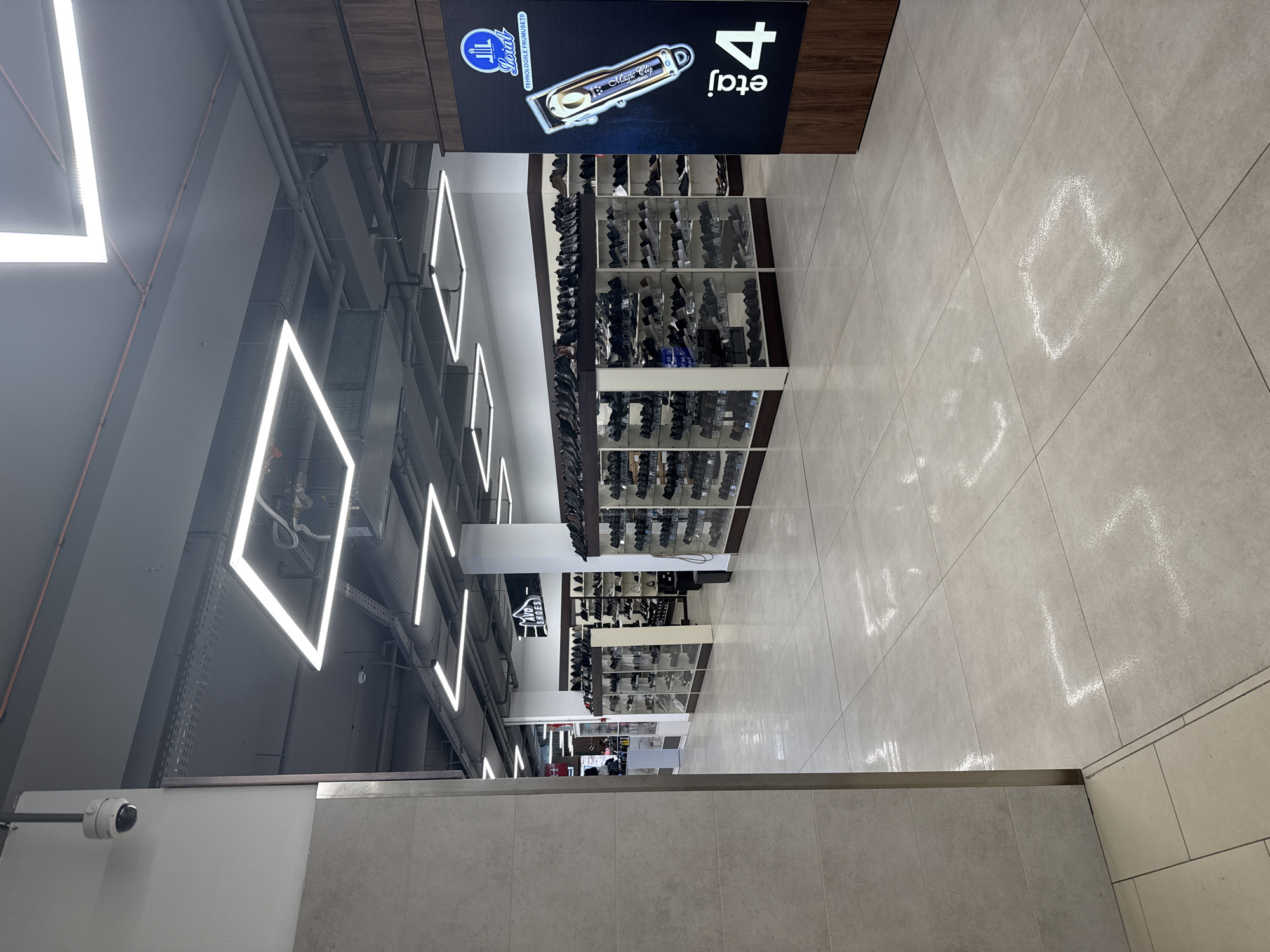 The image depicts the interior of a retail establishment, likely a shoe store, located in Chișinău, Moldova. The space is well-lit with modern rectangular fluorescent lighting fixtures. The flooring consists of large, light-colored tiles. In the foreground, a cream-colored wall with horizontal texturing is visible on the left.
Dominating the mid-ground are display shelves filled with shoes. These shelves are organized in rows and are made of glass and light-colored wood. The shoes are predominantly dark-colored, suggesting a variety of styles for men, women, and possibly children.
On the right side of the image, a prominent dark display board with wooden trim features a logo with text that appears to be "Loral" and the tagline "Tehnologiile Frumuseti" (Technologies of Beauty). Below this is an illustration of a hair clipper, and further down, the text "etaj 4" is displayed, indicating the fourth floor.
In the background, more shoe displays can be seen, along with a sign for "AVO SHOES" with a stylized shoe logo. A security camera is visible mounted on the ceiling in the upper left quadrant of the image. The overall impression is that of a modern, clean, and organized retail environment. There are no individuals clearly visible in the image.