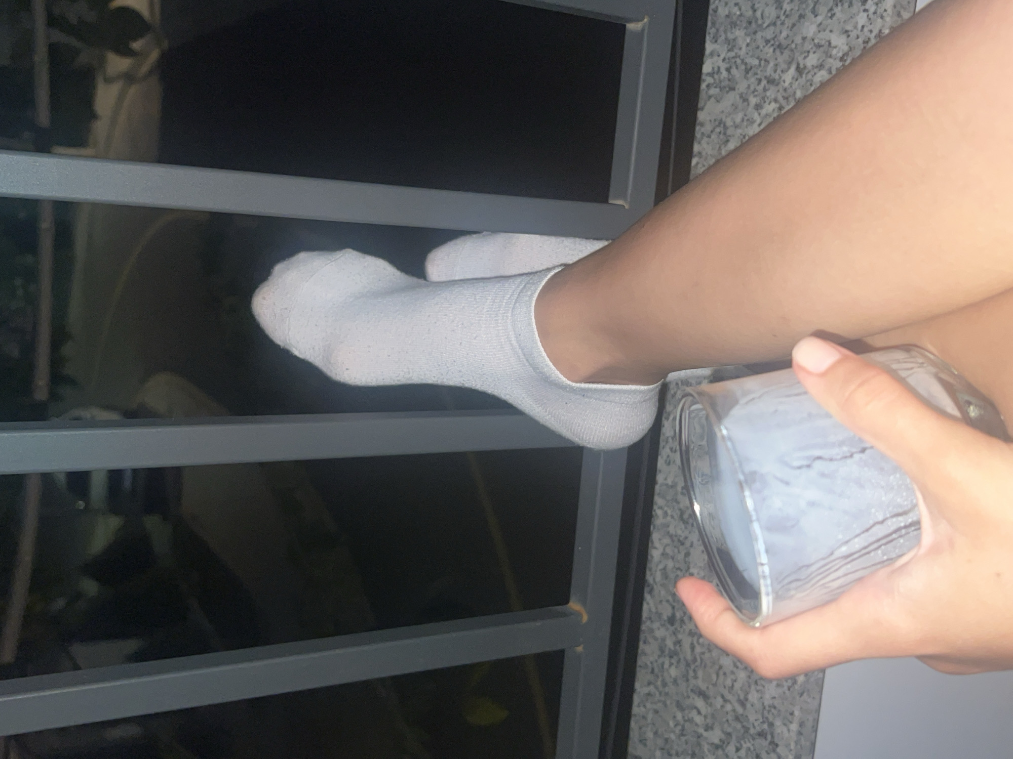 A close-up shot captures a person's leg clad in a white sock resting on a gray metal bar, with a hand holding a clear glass, possibly containing liquid, at the edge of the frame. The background is blurred, showing hints of greenery through the bars. The setting seems to be an outdoor space.