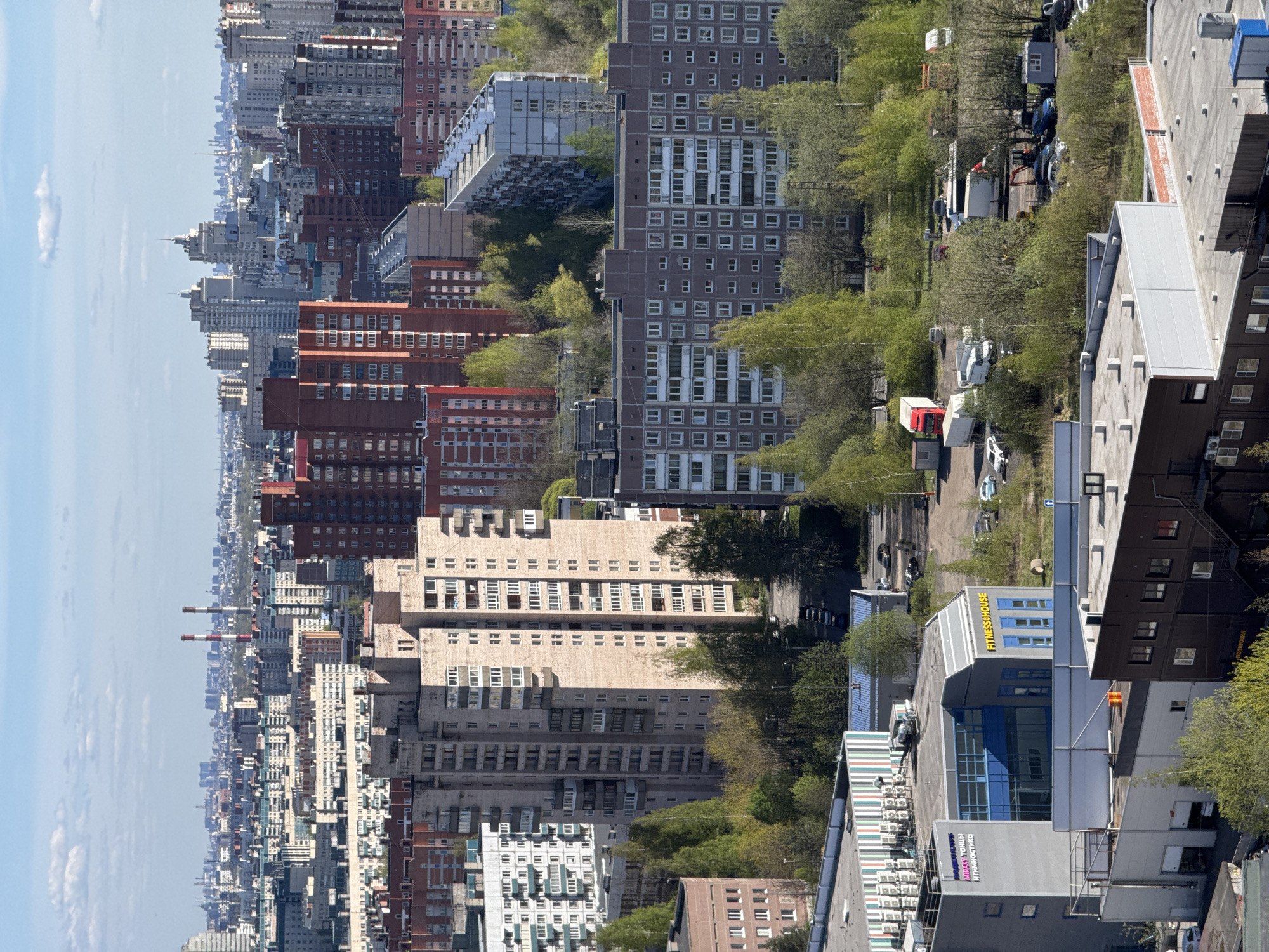 This image depicts a high-angle view of a cityscape, likely in Saint Petersburg, Russia, characterized by dense urban development and diverse architectural styles. The perspective includes a cluster of residential and commercial buildings varying in height, color, and age, interspersed with greenery. The architectural styles range from older, possibly Soviet-era structures to modern complexes. Various signs are visible on buildings, some in Cyrillic script. In the background, a hazy skyline suggests distance. The lighting indicates a bright, possibly sunny day. The overall impression is that of a densely populated urban area with a mix of old and new structures.
