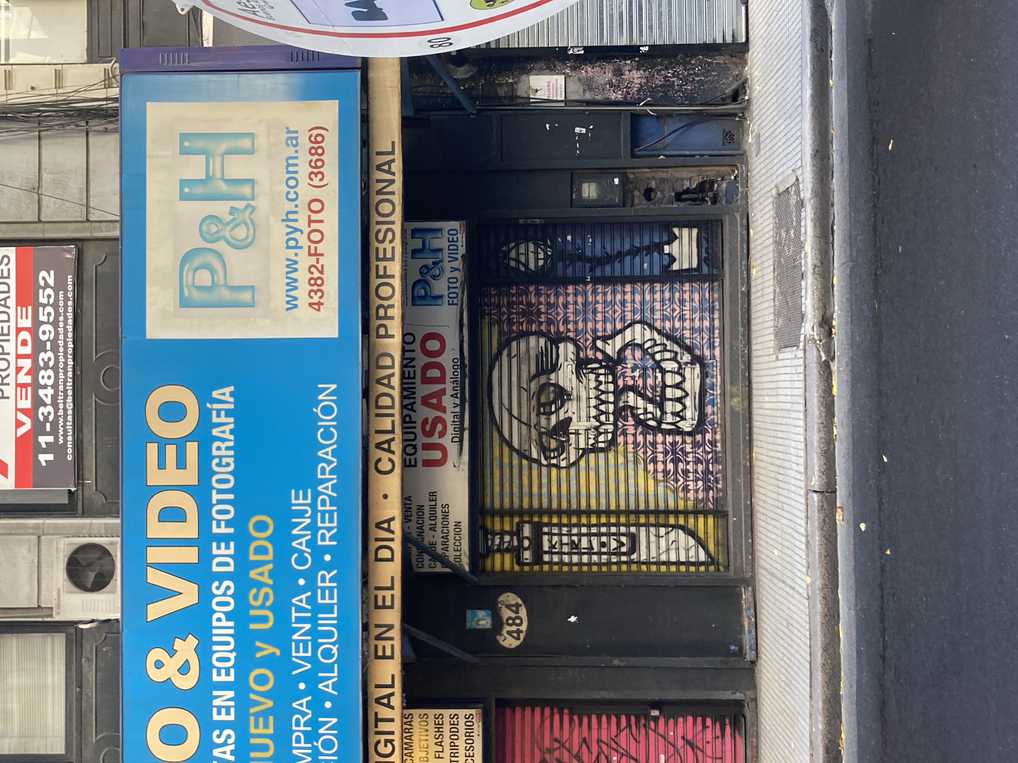 The image is an exterior shot of a business in Buenos Aires, Argentina. The business, "P&H," specializes in photography and video equipment. The storefront is partially covered by a metal rolling gate that is adorned with graffiti, including a skull. The address, 484, is visible on the facade. The sign indicates that they buy, sell, and repair new and used equipment, and offer digital and analog photography supplies. The sign also lists their phone number (4382-FOTO (3686)) and website (www.pyh.com.ar). Beside this shop is a place for selling properties.