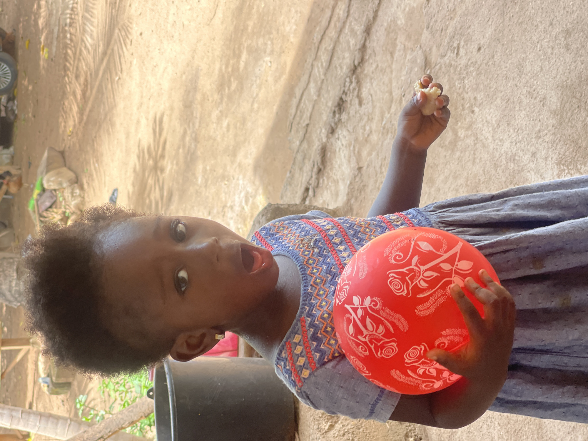 A young child, likely a girl, with dark skin and short, curly dark hair is holding a red balloon with white rose designs in her right hand and a piece of food in her left hand. She is wearing a blue dress with a patterned yoke. Her mouth is open in what appears to be an expression of surprise or excitement. A black bucket is visible near her. The background consists of a sunlit, sandy or earthen area with vegetation and what looks like parts of a vehicle. Without further information, it is impossible to confirm the location in the city of Akwanga, Nigeria.