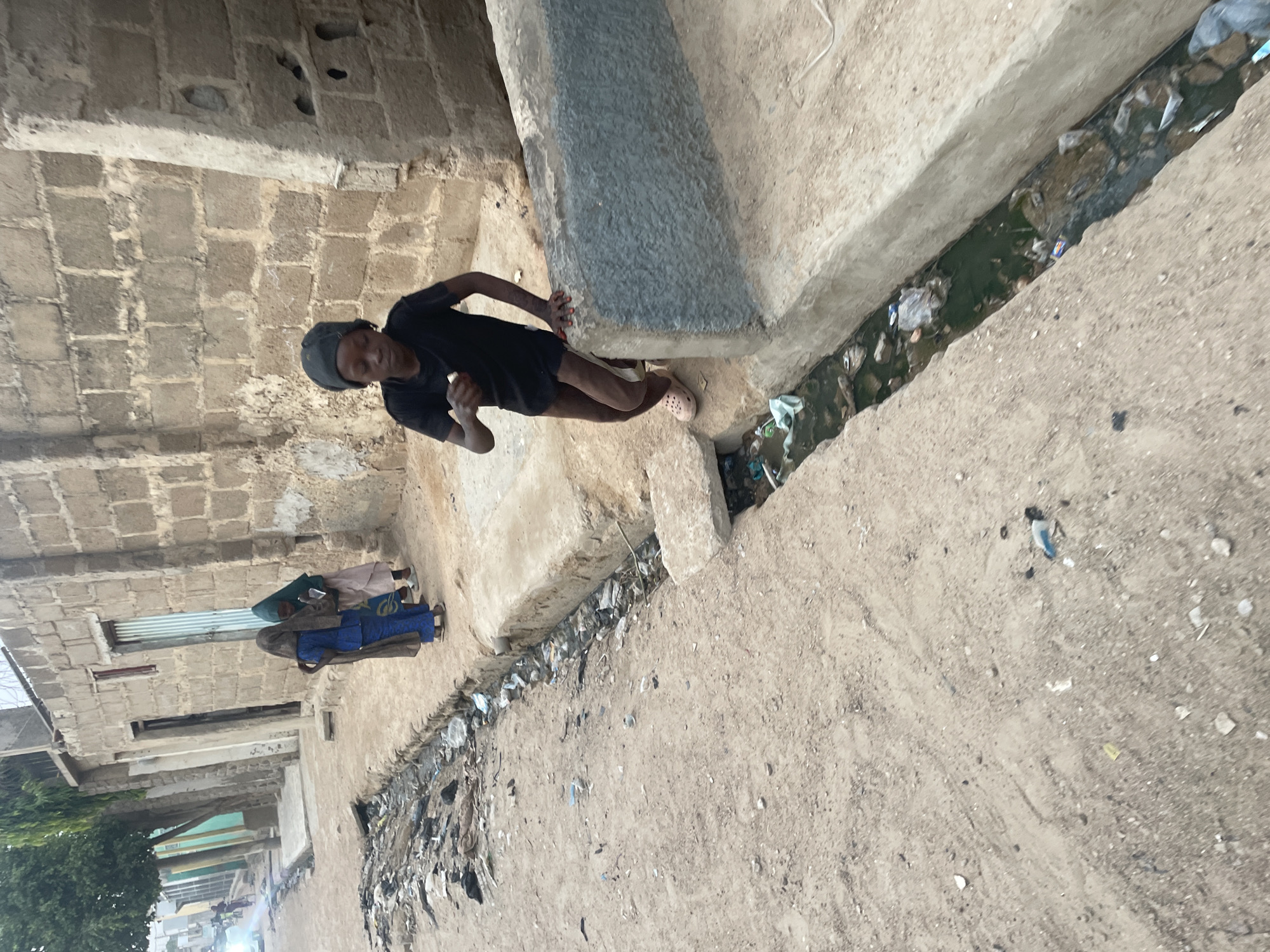 The image depicts an individual in a black t-shirt and a cap, standing next to a concrete structure, possibly a part of a building or infrastructure. They appear to be holding something in their hand. A drainage ditch filled with debris and stagnant water runs alongside a dusty street or pathway. In the background, there is a building with block construction, and two individuals are present, covered with clothing. The location is in a built-up area with visible signs of litter and urban degradation. The image does not provide specific clues about the exact location in the city of Unknown, Unknown.