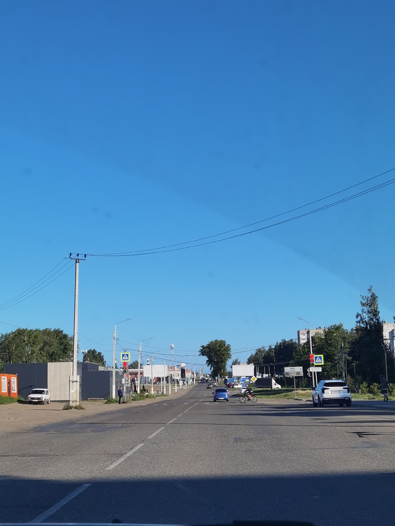 A sunny, clear blue sky is visible above a straight, paved road in Chaikovsky, Russia. The road has a pedestrian crossing marked with white lines, and traffic signals are present at an intersection. On the left side of the road, there is a white fence and a small orange building. Further down the road, several cars are seen, including a white SUV and a blue sedan. A person is riding a bicycle in the middle of the road. Trees and buildings line the sides of the road, with power lines stretching across the scene. The overall impression is a typical day in a Russian town.