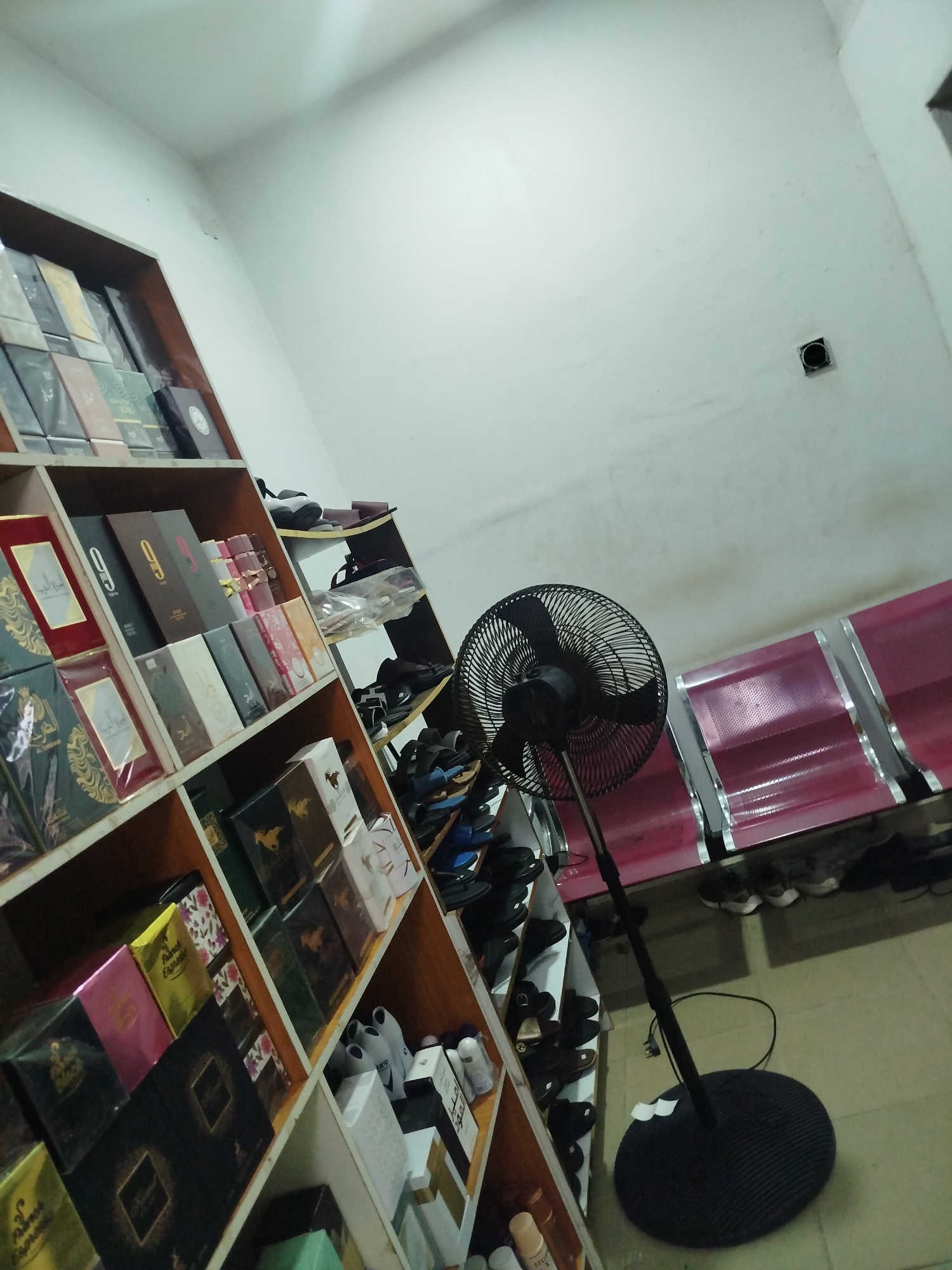 The image depicts a retail space in Uselu, Nigeria, showcasing an array of boxed products, primarily perfumes and colognes, arranged on wooden shelves. The products are diverse in packaging, with some featuring elaborate designs and others simpler, modern aesthetics. A black oscillating fan stands on the floor in the foreground, angled towards the shelves. In the background, a row of red and silver seating suggests a waiting area. Scattered on the floor and lower shelves are various pairs of footwear, including sandals and shoes, indicating a busy environment. The walls are painted white, and the lighting appears to be artificial, casting a neutral tone over the scene. The overall impression is of a well-stocked store, likely a perfumery or a general store with a significant fragrance section.