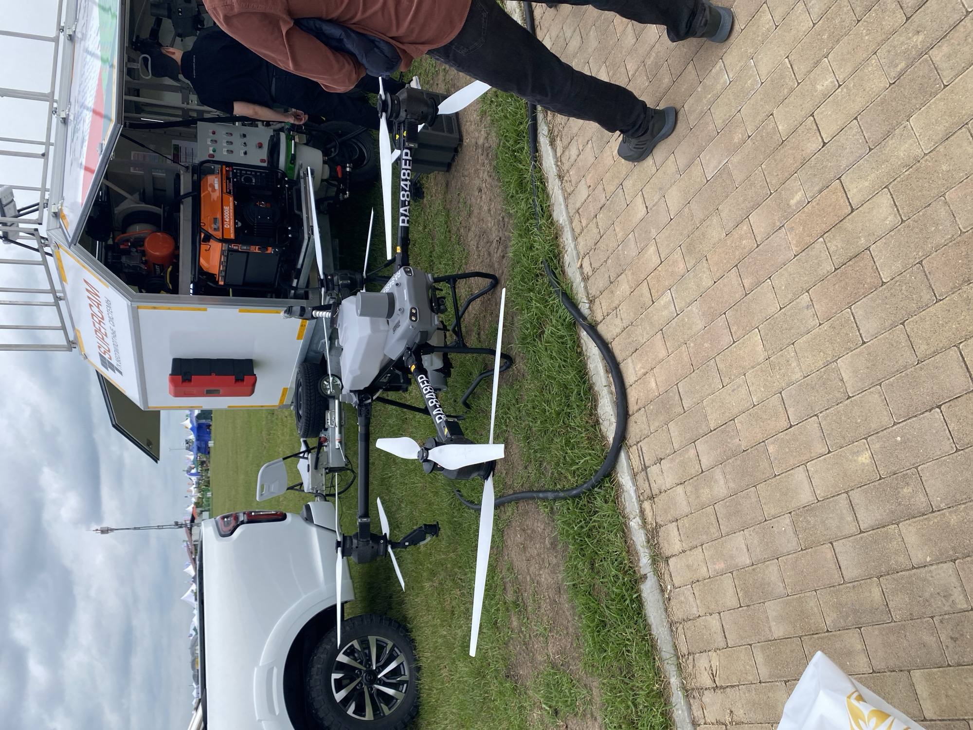 The scene is outdoors. There's a drone on grass near a brick walkway. Two individuals are present; one is partially visible wearing a brown jacket and black pants and the other one is partially visible wearing a black jacket. A white vehicle with a trailer is parked nearby. The trailer contains orange and black equipment. The drone is gray with white blades and has the text "RA-848EP" on its arm. The image was possibly taken at Voronezhskaya, Russia.