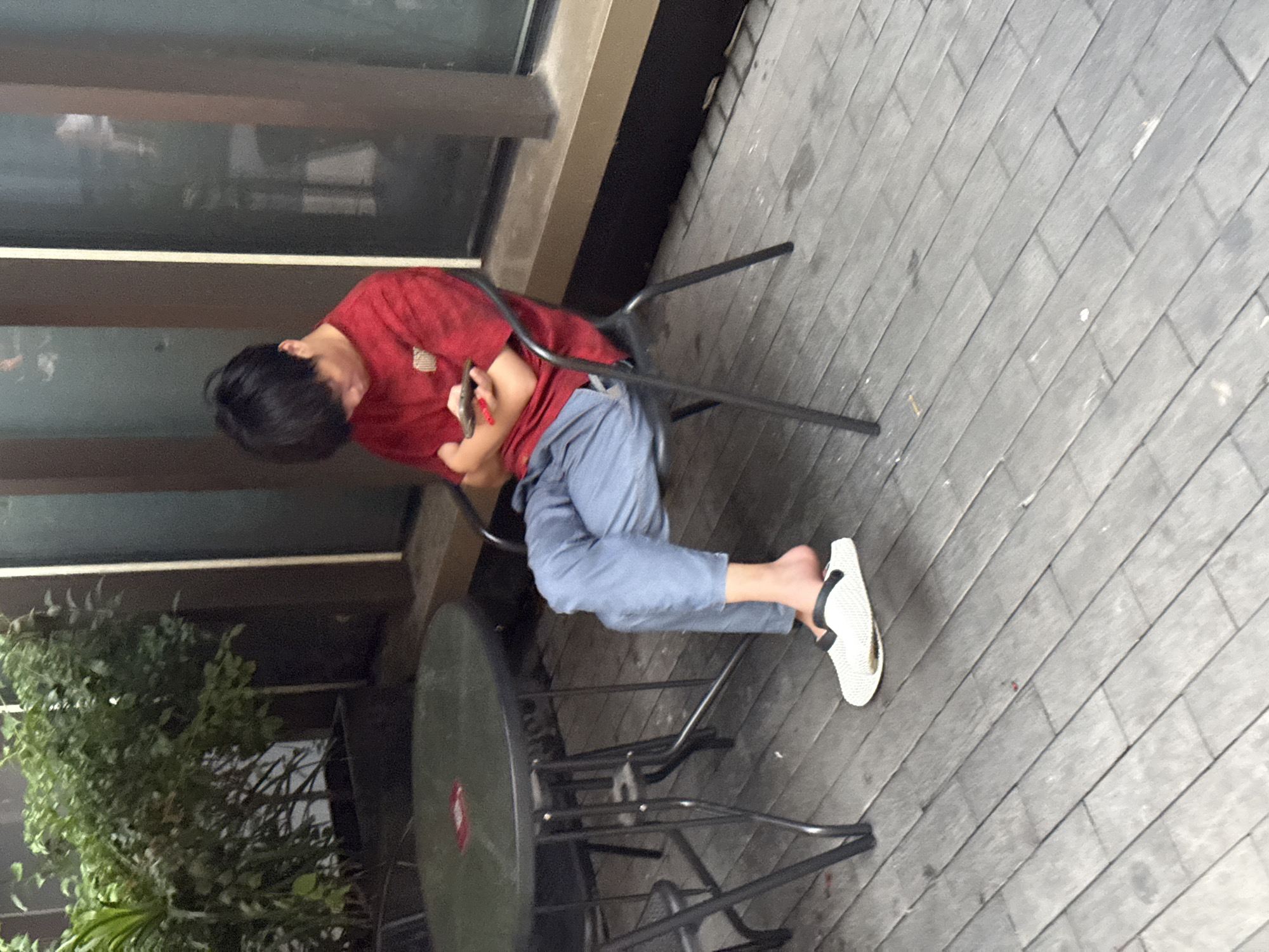 A young person with dark hair is sitting on a black metal chair at an outdoor cafe. They are wearing a red t-shirt with a graphic design and light blue pants. Their left foot is bare except for a white slide sandal with a black strap, and their right foot is also bare. The person is looking down at a mobile phone held in their hands. A round, dark green outdoor table with metal legs is to their left. The cafe seating area is on a wooden deck. Behind the table, there is a planter box with lush green plants. The background shows a building facade with windows and vertical brown and dark brown elements. The scene is set in Yuzhong District, China.