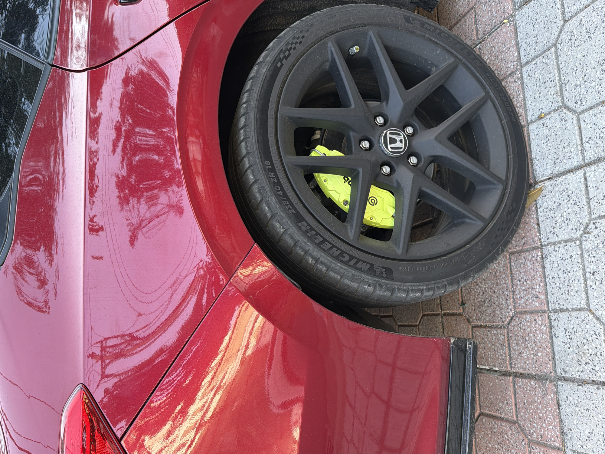 The image shows a close-up view of the rear wheel and fender of a red car, parked on a tiled pedestrian walkway. The car has a glossy finish that reflects the surrounding environment, including power lines and trees, suggesting an outdoor urban setting. The wheel is black with a bright yellow brake caliper visible behind the spokes, a detail often associated with performance vehicles. The tire is a Michelin Pilot Sport 4. The ground is paved with interlocking hexagonal tiles in a pattern of pink, white, and grey. The car appears to be a Honda, indicated by the emblem on the wheel. The lighting suggests it is daytime. The scene is static, with no people or activity depicted. The location is identified as Nha Trang, Vietnam.