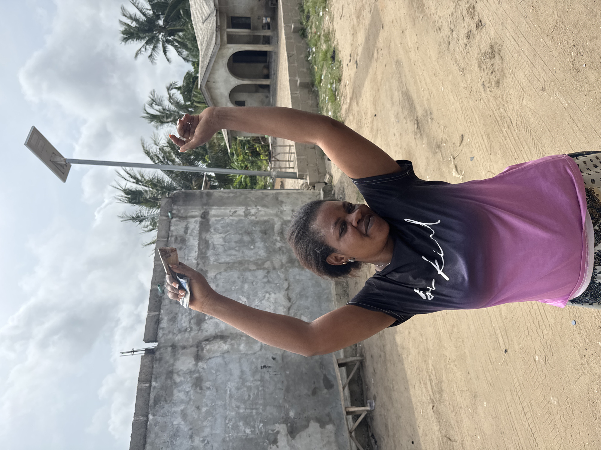 A woman stands at an angle, smiling, and holding money in her right hand. She is wearing a dark blue t-shirt with a light purple section and patterned bottoms. The background shows a dusty road and buildings. To the left of the woman is a grey concrete building with a solar panel on a pole. Behind the woman, there is a building with arched openings and palm trees. The image is located in Badagry, Nigeria.