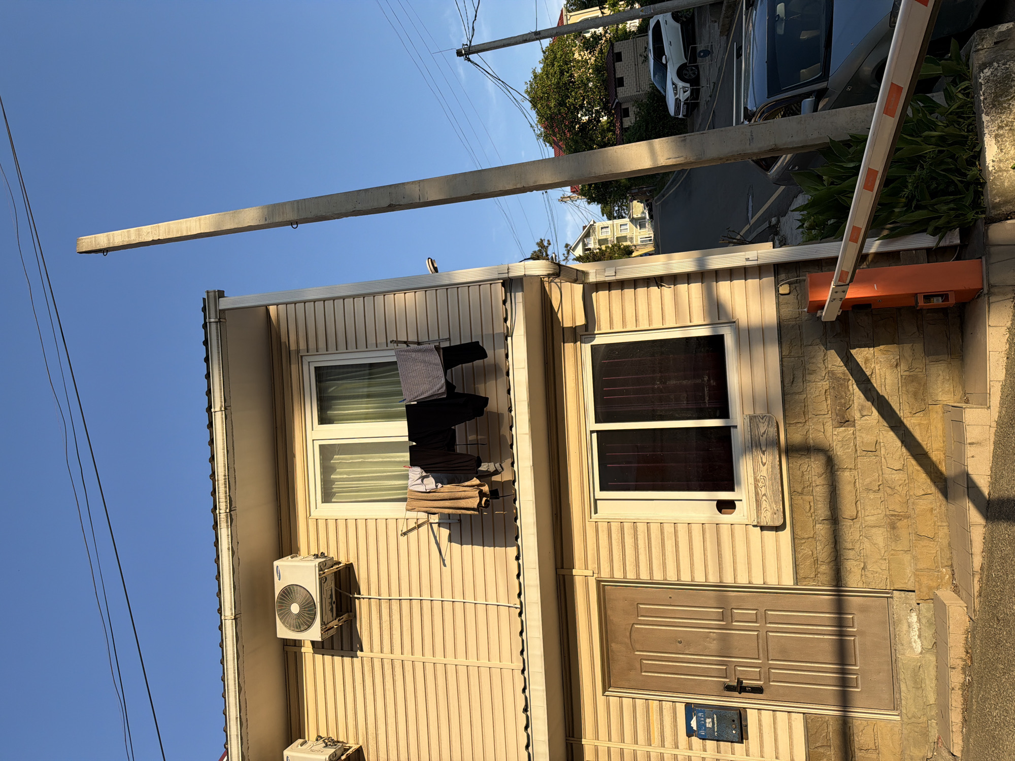 The image captures a view of a two-story building with beige siding and a brown door. Clothes are hanging on a line outside a second-story window. Two air conditioning units are mounted on the exterior wall. A concrete utility pole stands tall in the foreground, leaning slightly. Beyond the building, a street winds uphill, with parked cars and other buildings visible. Trees and foliage are scattered throughout the scene. The sky is clear and blue, with the sun casting long shadows, suggesting it is late afternoon or early evening. This scene is characteristic of a residential area in the city of Sochi, Russia, a popular resort town on the Black Sea coast.