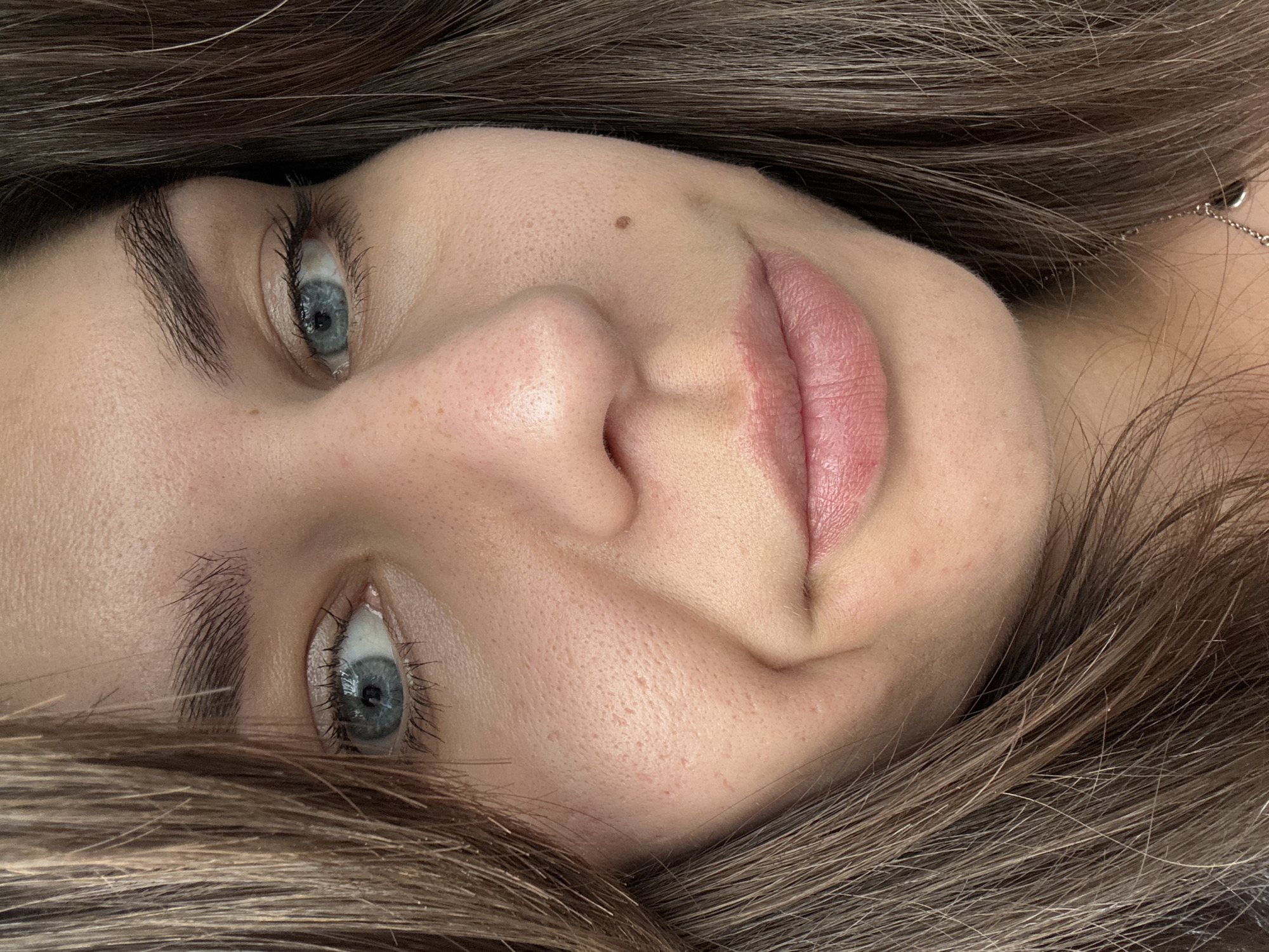 A close-up, over-the-shoulder shot of a person's face from the nose up. The subject has light skin with visible pores, some freckles, and a small brown mole on the right cheek. Their eyes are open and blue, with dark lashes and eyebrows. The lips are slightly parted and appear to be wearing a light pink lipstick. The person's brown hair frames their face. The background is out of focus and appears to be indoors. The image does not provide any information about the location within a city.