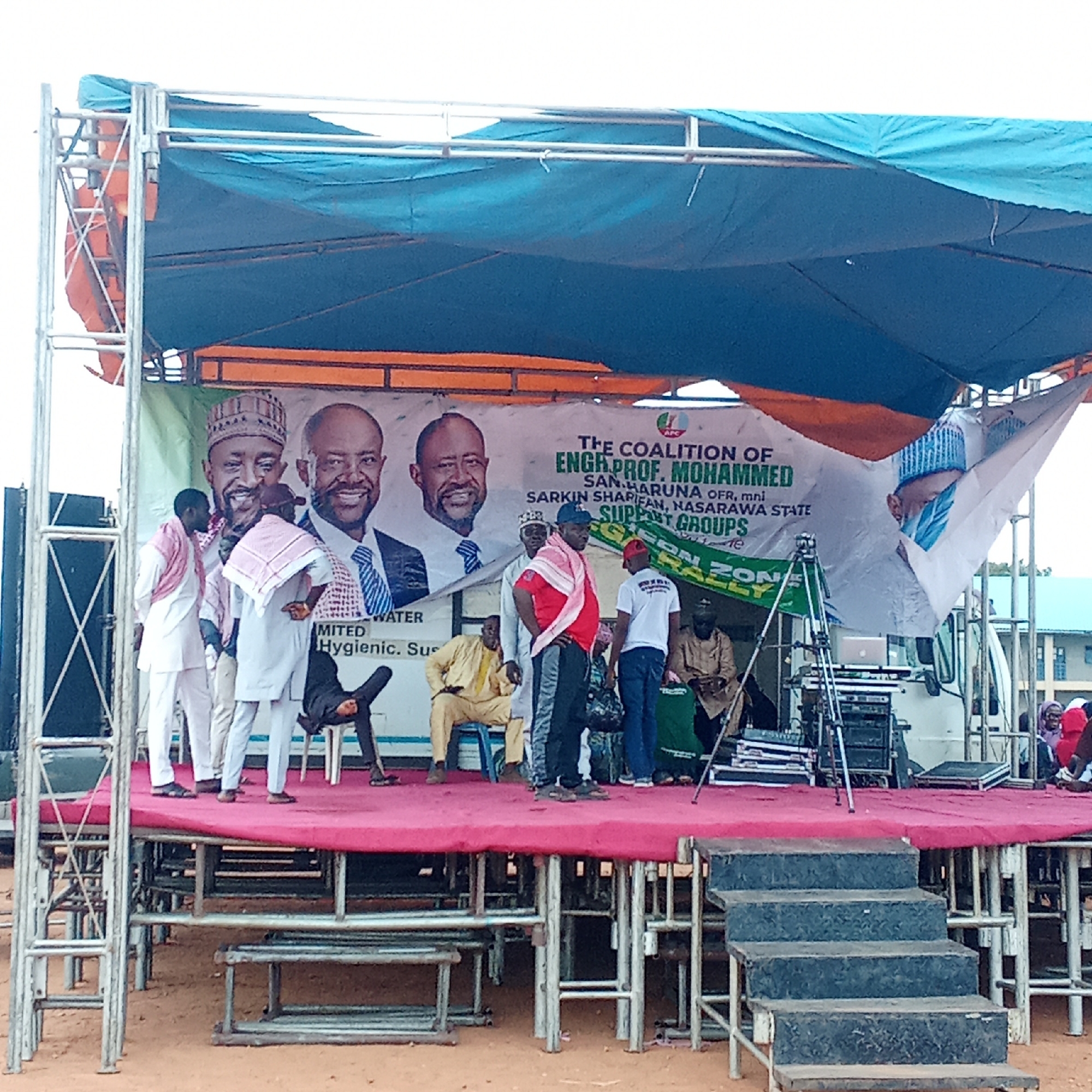 The image shows a stage set up in Akwanga, Nigeria. The stage is covered with a red carpet and has a backdrop banner featuring portraits of Engr. Prof. Mohammed San Haruna, with the text "THE COALITION OF ENGH PROF. MOHAMMED SAN HARUNA OFR, mni SARKIN SHARIFAN, NASARAWA STATE SUPPORT GROUPS." Several people are on the stage, including some dressed in traditional Nigerian clothing. A man is seen leaning backwards. There are also audio equipment and other supplies on the stage, and a white truck is visible in the background. The stage is made of scaffolding and metal structures, with steps leading up to it. The ground is sandy.