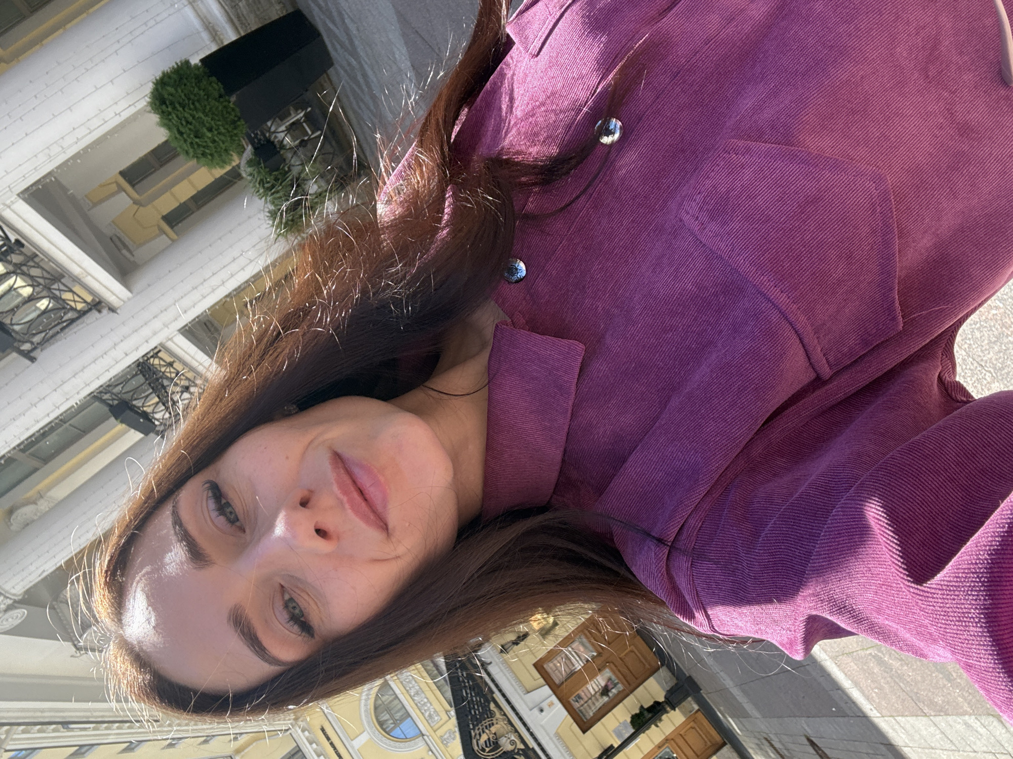 A woman takes a selfie outdoors in front of a building. She has long brown hair and is wearing a purple corduroy jacket. The background shows a building with classical architecture and a potted plant. The overall lighting suggests it is daytime, with sunlight casting shadows. The image appears to be taken on a street in Saint Petersburg, Russia, based on the architectural style of the building.