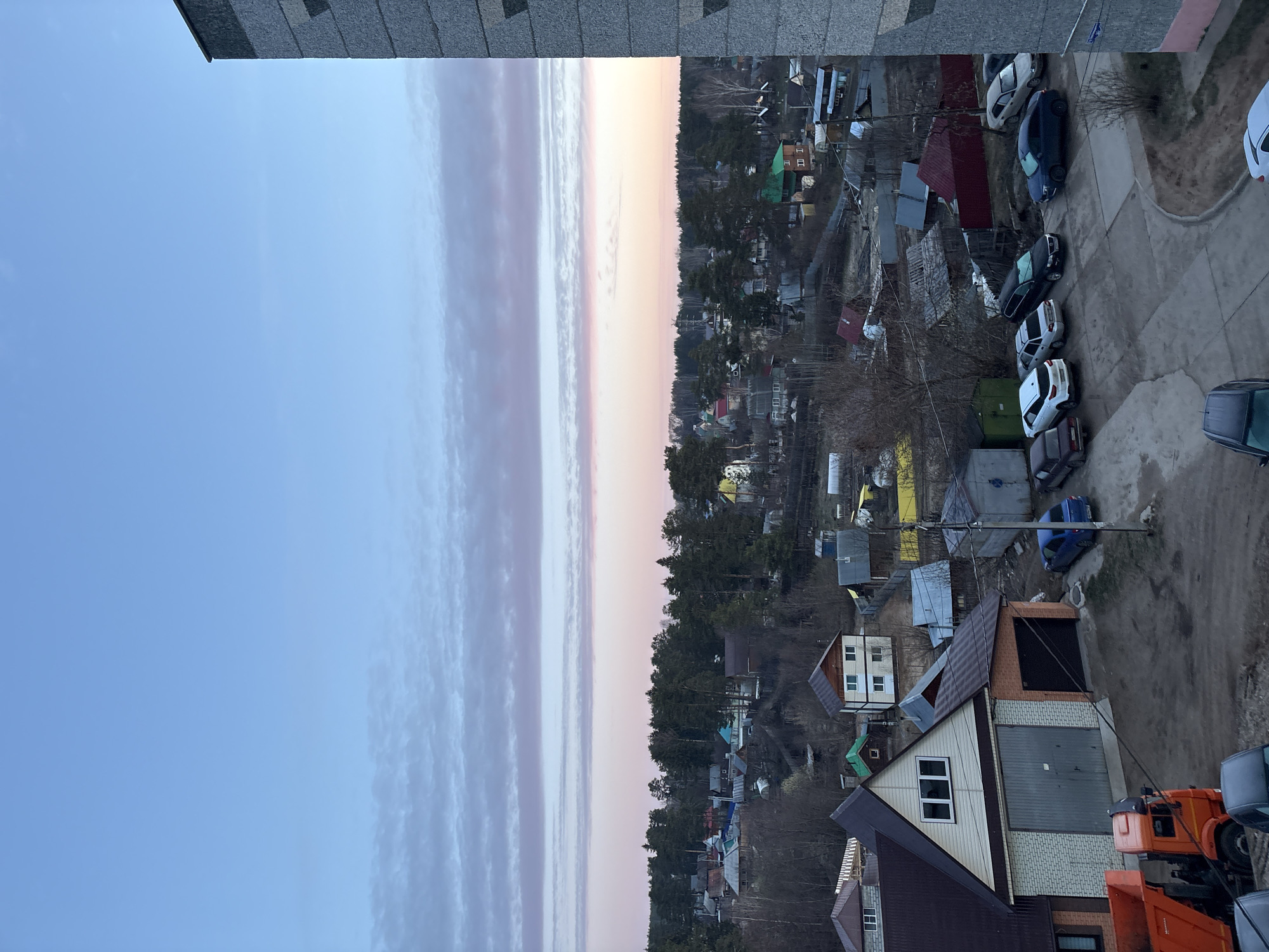 An elevated view shows a residential area in Chaikovsky, Russia. The sky transitions from blue to pink, with layered clouds. Below, dense trees separate residential buildings, varying in size and color. A row of parked cars lines a concrete area. At the bottom, parts of a house, a garage, and a truck are visible. No people are observed.
