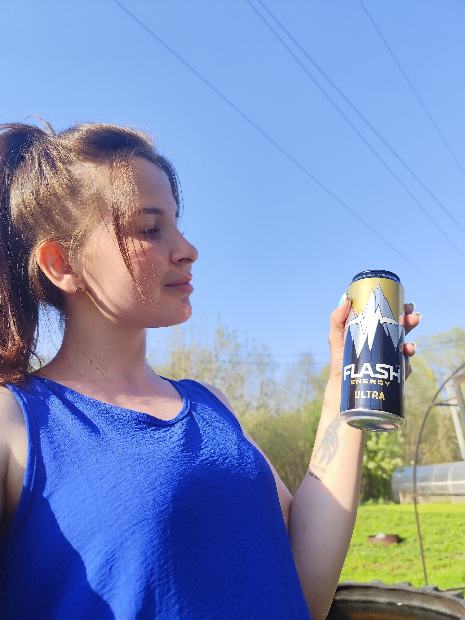 A young woman with light brown hair pulled back in a ponytail is holding a can of Flash Energy Ultra. She is wearing a blue sleeveless top and is standing outside on a sunny day. The sky is clear blue, and power lines are visible in the background. Behind her, there is green grass, trees, and a greenhouse. There's a tire on the ground near her. The woman is looking at the energy drink can. The image appears to have been taken in Фоки, Russia.
