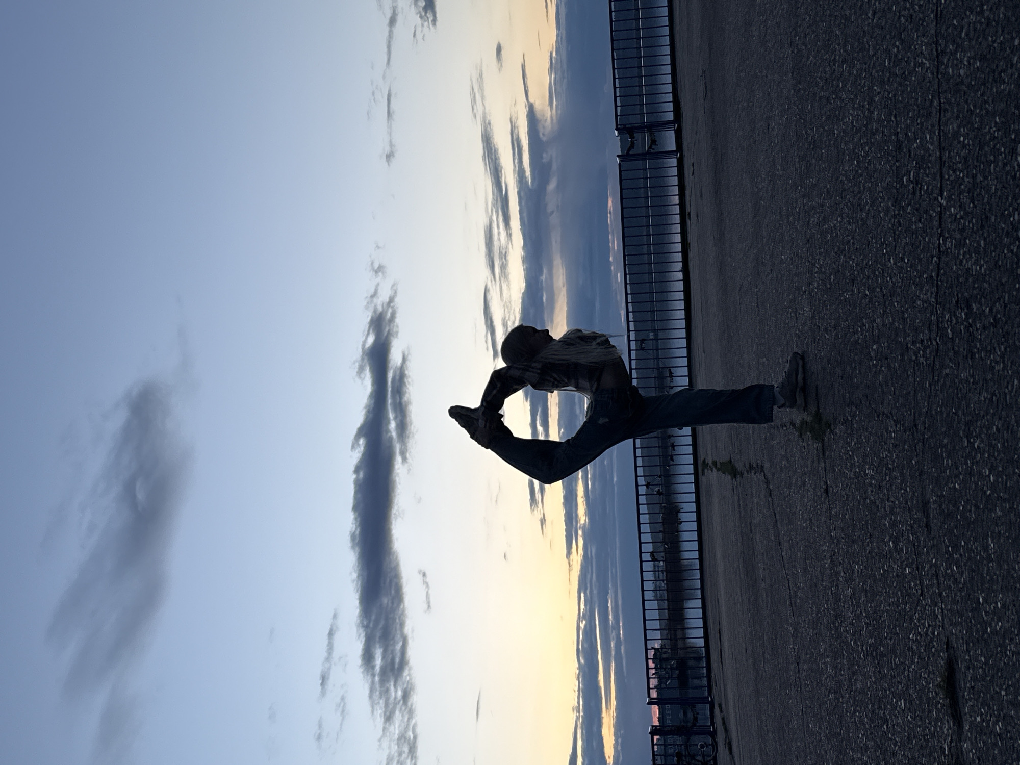 The image displays a full-body silhouette of a person performing a complex acrobatic or dance pose against a twilight sky. The individual, positioned slightly to the left of center, is captured in motion with their body arched back and one leg extended high towards their head, forming a "needle" or standing split. They appear to have light-colored hair and are dressed in a plaid shirt and jeans.
The background is dominated by a sky exhibiting the soft colors of either sunrise or sunset, transitioning from light blue at the top to warmer yellow and orange hues closer to the horizon. Scattered clouds are visible across the sky. To the right, a dark metal fence or railing runs along the edge of a paved, dark-textured surface, likely asphalt or concrete, which occupies the lower portion of the frame. Beyond the fence, an indistinct body of water or distant landscape can be seen, with subtle dark outlines of what might be structures on the far horizon. The overall lighting creates a strong silhouette effect, with the person and foreground elements appearing dark against the brighter sky. No text is visible in the image.