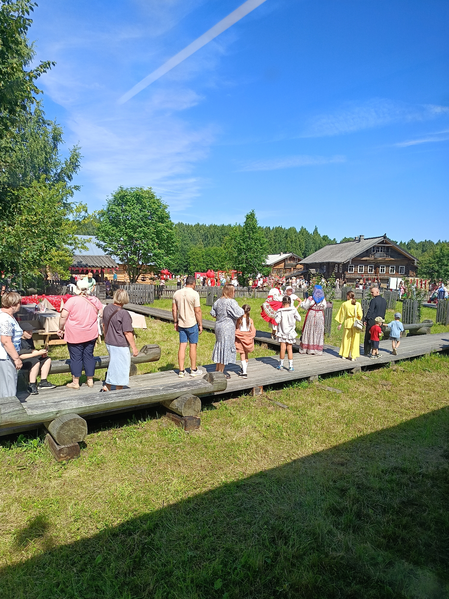 In an outdoor, rural setting under a clear blue sky with a contrail, a wooden walkway constructed from large logs and planks extends diagonally across the frame. The walkway is populated by a group of people, some dressed in traditional clothing. To the left, a grassy area features picnic tables with red and white checkered tablecloths. In the background, several rustic wooden buildings with dark roofs are visible, characteristic of a village or historical reenactment site. The overall scene depicts a sunny day at a cultural event.
