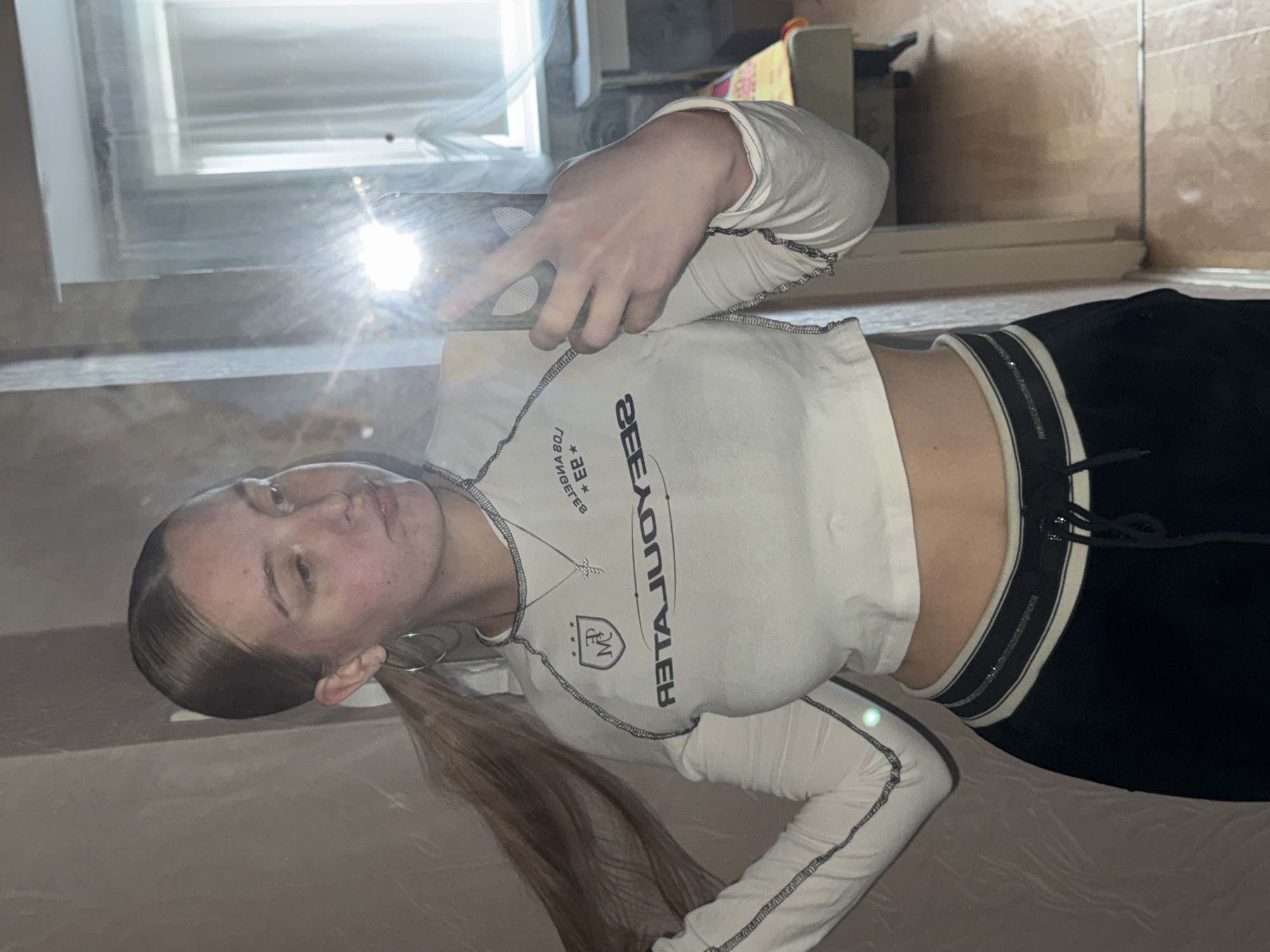 A young woman is taking a mirror selfie in an indoor setting. She has blonde hair tied in a ponytail and is wearing a white, cropped long-sleeved shirt with black trim and text that appears to be "LOS ANGELES" and "BEAUTIFUL". She is also wearing black pants with a striped waistband. Her right hand is holding a phone, and her left hand is touching her hair. The background shows a wall, a mirror reflecting the scene, and a window with sheer curtains. There is also a glimpse of a bed with a yellow and red patterned cover in the background. The environment suggests a residential interior. The specific location in the city of Perm, Russia, cannot be determined from the visual information provided.