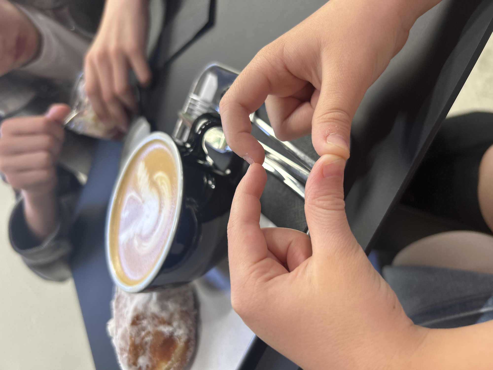 This is a medium shot, eye-level, interior photograph showing a table setting with a donut and coffee. The focus is on two hands in the foreground, forming a heart shape with their fingers. To the left, a sugar-dusted donut sits on a white plate. To the right, a black mug filled with latte art coffee is present. In the blurred background, two individuals are visible; one is stirring something in a small container with a spoon, and the other appears to be looking at their hands. The lighting is natural and comes from the left. The scene is set in a cafe or restaurant in Izhevsk, Russia.