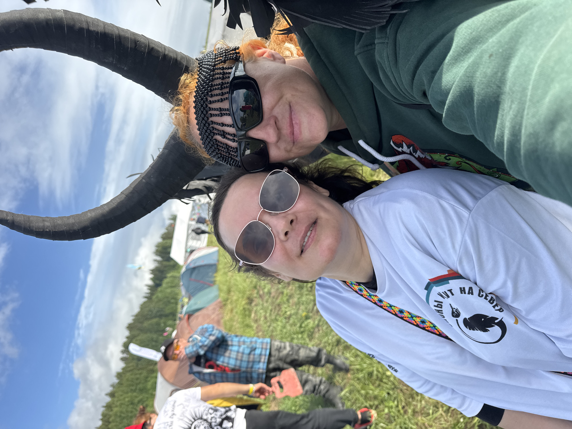 This image captures a selfie of two women at an outdoor event or festival in Большая Кивара, Russia, under a bright blue sky with scattered white clouds. The photo is taken from a slightly upward angle, showcasing a grassy field and distant trees.
In the immediate foreground, two women are the primary subjects. The woman closer to the camera has dark hair and wears light-framed, round sunglasses, offering a faint smile. She is dressed in a white long-sleeved shirt featuring Russian text that reads "ИДУТ НА СЕВЕР" (IDUT NA SEVER, translating to "GO NORTH" or "THEY GO NORTH"), accompanied by a graphic of a feather within a circular emblem. A colorful, intricately patterned strap is visible across her chest. Behind her, the second woman has reddish-blonde hair, sports dark, rectangular sunglasses, and a distinctive black beaded headpiece. She wears a dark green hooded sweatshirt and maintains a neutral expression.
A large, dark, curved object, resembling a horn or an artistic prop, partially frames the upper left corner of the image. The background reveals a lively outdoor scene with several tents pitched on the grassy ground, suggesting a campsite or festival setting. Two other individuals are visible relaxing on the grass; one person in a blue plaid shirt and dark pants is lying on their back, and another, partly obscured, wears a light top with a dark graphic and dark pants. The clear daytime lighting and relaxed poses of the subjects contribute to a convivial and casual atmosphere at this outdoor gathering.