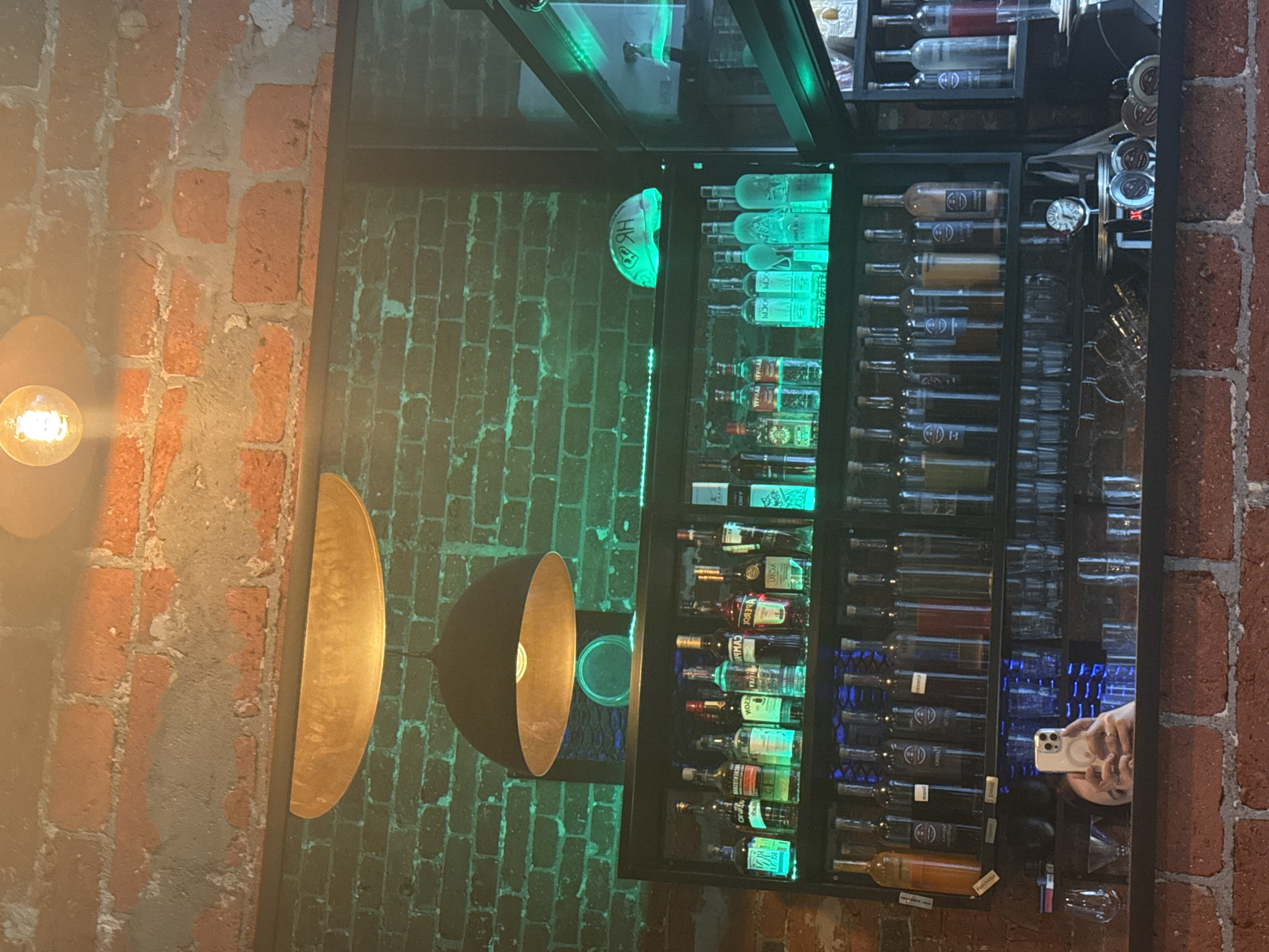 The image shows a bar area with a dark-framed liquor cabinet filled with various bottles illuminated with green and blue lights. The backdrop includes brick walls on both sides of the cabinet. A person is taking a picture with a white smartphone, their reflection visible in the cabinet's glass door. Two wall-mounted lamps with golden interiors are positioned on the left. The location appears to be Moscow, Russia, based on Cyrillic lettering on some bottles.